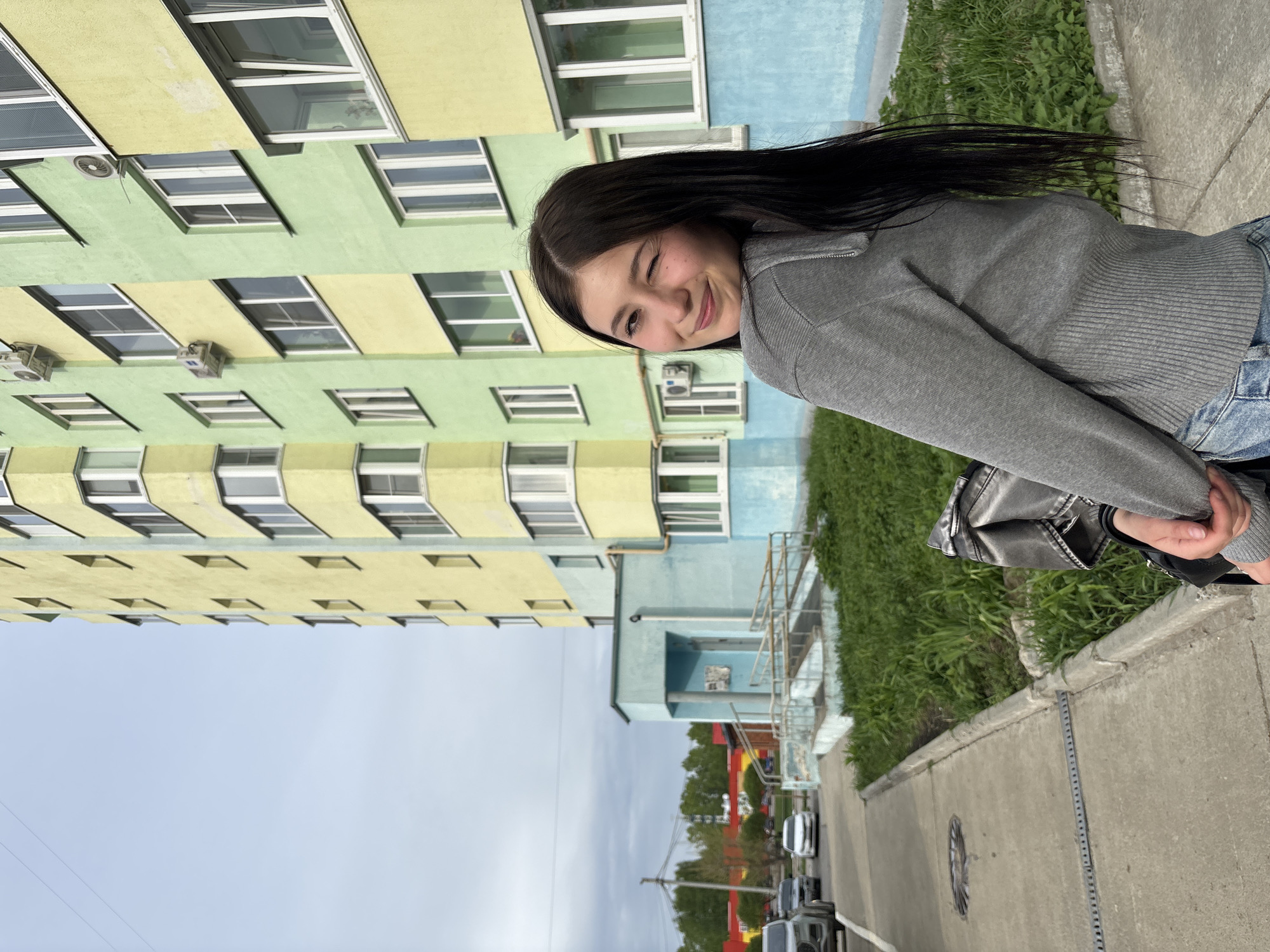 A young woman with long, dark hair, wearing a grey sweater and blue jeans, stands on a sidewalk and winks at the camera. Behind her is a large, multi-story apartment building with windows in a repetitive pattern. The building is painted in alternating bands of green, yellow, and blue. A street with cars and green trees stretches into the distance. This location is in the city of Chaikovsky, Russia.