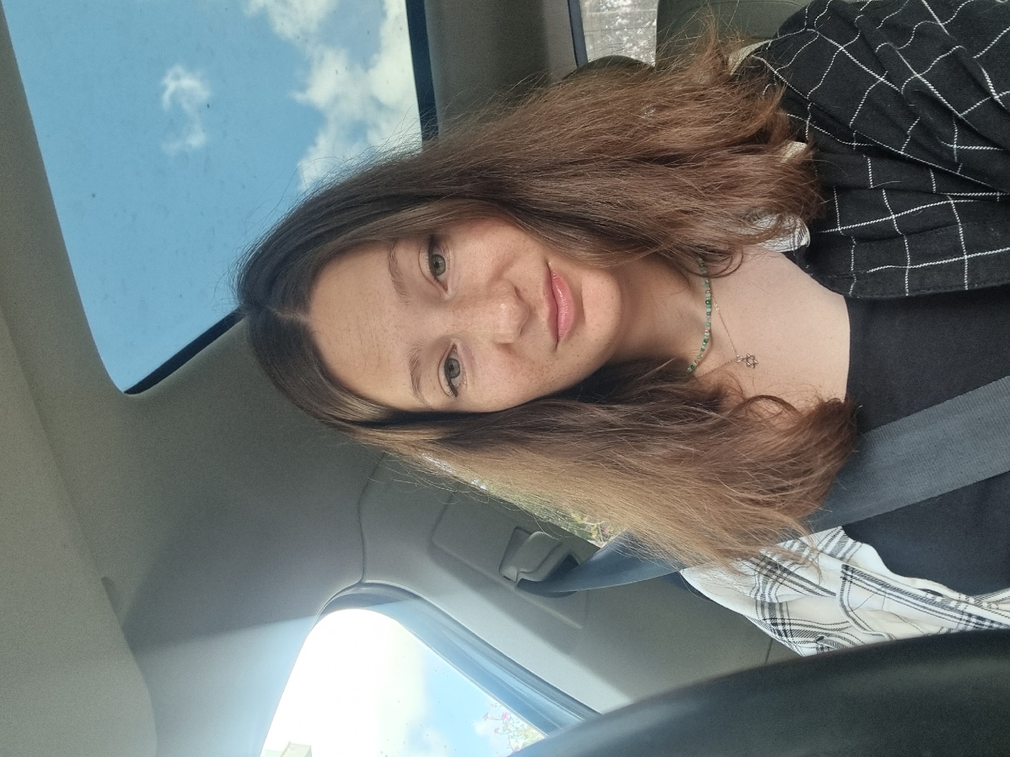 A person is seated in a car. The person has light brown hair and freckles. They are wearing a black shirt, a black and white plaid shirt, and a seatbelt. They also have a necklace with a Star of David pendant. The car has a sunroof. The scene appears to be in a car, based on the interior elements visible. No additional information is available to determine if the photo was taken in Ramat Gan.