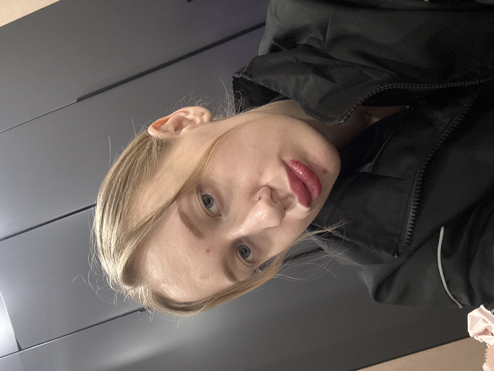 A close-up, rotated selfie photograph shows a woman with blond hair, light skin, and blue eyes. She is wearing a black jacket. Her lips are colored with a dark lipstick. The background appears to be a gray wall or cabinet. The image lacks sufficient information to determine the location in Novoaleksandrovka, Russia, or to provide any additional contextual details.