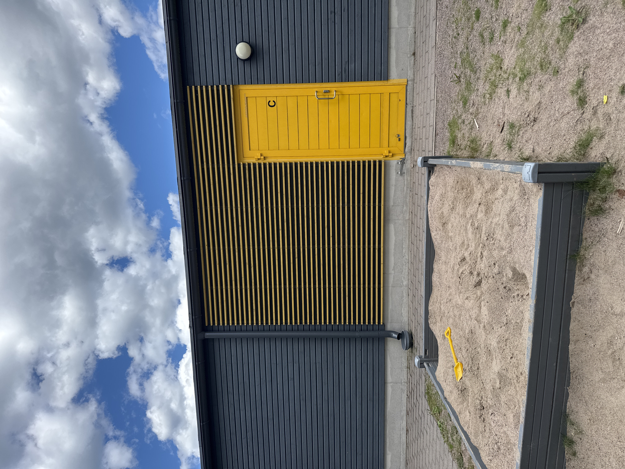 This is a medium shot, outdoor photograph of a modern building with a bright yellow door and a sandbox. The building is clad in dark gray vertical planks, with a section above the door featuring yellow vertical slats. A white circular light fixture is mounted on the upper part of the dark gray wall. The yellow door has a metal handle and a small lock mechanism. To the right of the building, a sandbox filled with light brown sand is visible. A small yellow shovel rests in the sand. The ground surrounding the sandbox is a mixture of sand, gravel, and sparse green grass. A gray pipe with a curved end extends from the building down towards the ground. In the background, a cloudy blue sky is visible. The photograph is taken from a slightly elevated angle.