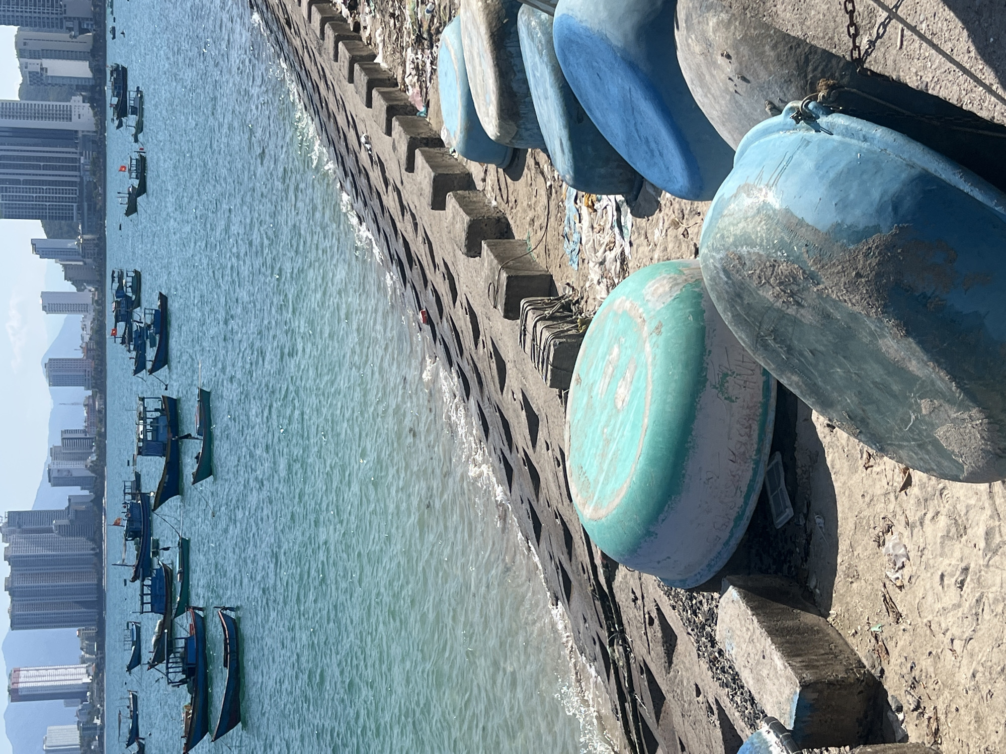 This image depicts a coastal scene in Nha Trang, Vietnam, under a clear sky. In the immediate foreground, a sandy and rocky shore is visible, populated by approximately eight overturned traditional Vietnamese basket boats (thúng chai) of varying sizes, predominantly in shades of blue and faded turquoise-green. These boats exhibit signs of wear and weathering, with some showing attached ropes or chains. The ground around them contains scattered debris, including plastic fragments and fabric.
A concrete seawall or embankment runs horizontally across the mid-ground, separating the land from the water. This structure consists of dark gray interlocking concrete blocks arranged in a stepped pattern, with a top layer of rectangular blocks forming a border. Gentle waves lap against the lower section of this seawall.
Beyond the seawall, the turquoise-blue ocean extends, featuring calm water. Numerous fishing vessels are anchored or drifting in the water. These include various traditional wooden fishing boats, some with small cabins, and several smaller open motorboats. Approximately twenty-five boats are discernible, with some displaying red and yellow Vietnamese flags.
In the background, a dense urban skyline is visible, characterized by numerous high-rise apartment and hotel buildings, indicative of a developed coastal city. Distant mountains are faintly visible behind the city structures under a bright sky. No individuals are visible in the scene.