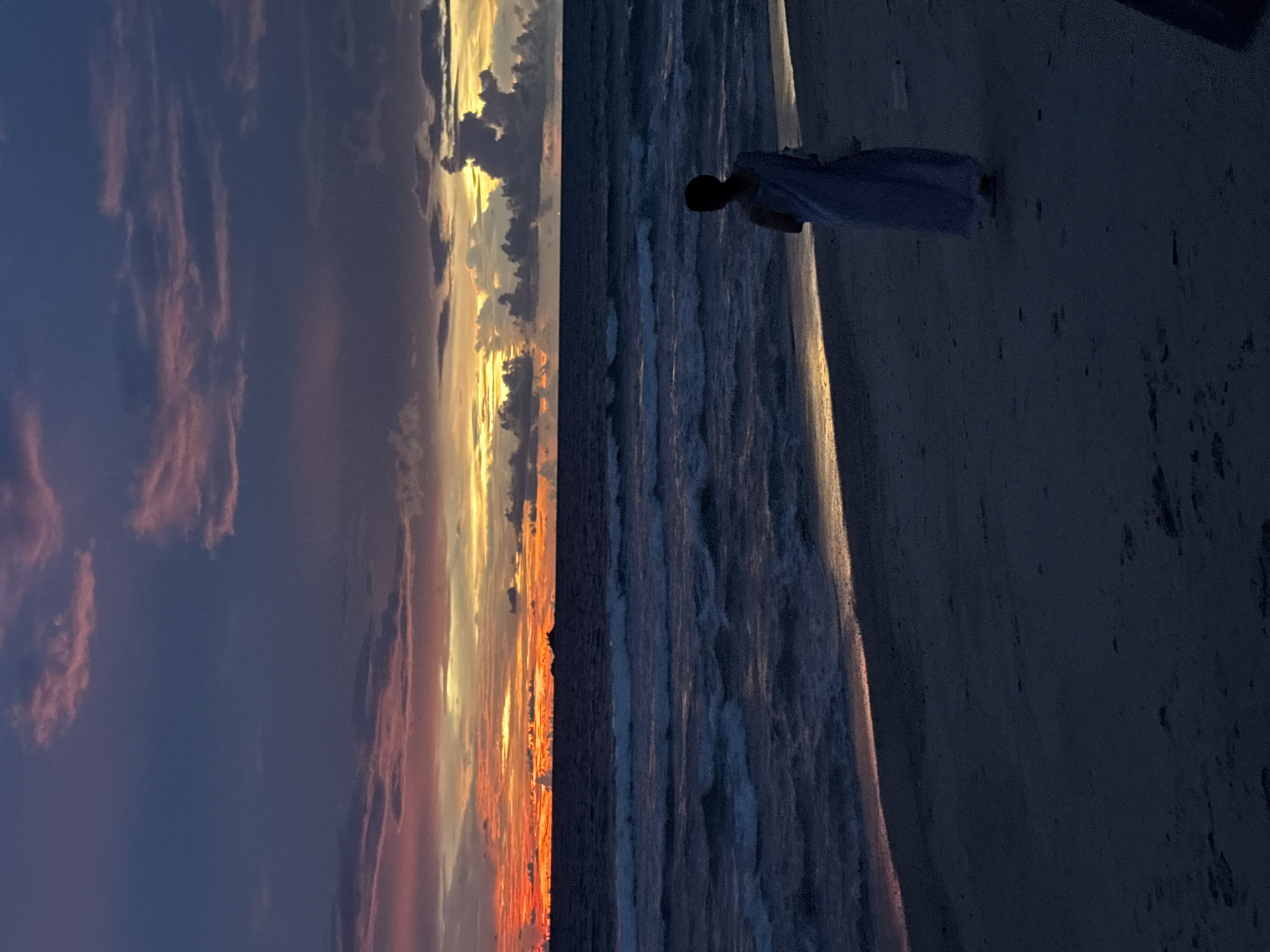 This is a vertical, nighttime photograph of a person standing on a beach in Choeng Thale, Thailand. The sky is filled with a dramatic sunset, with hues of orange, yellow, and purple painting the clouds. The sun appears to be setting over the ocean, casting a warm glow on the water. The waves are gently lapping at the shore, creating a peaceful and serene atmosphere. The person is silhouetted against the sunset, wearing a light-colored garment, and facing the ocean. The beach is sandy, with some scattered debris, and the water is dark, reflecting the colors of the sky. The overall mood of the image is tranquil and awe-inspiring, capturing the beauty of a tropical sunset.