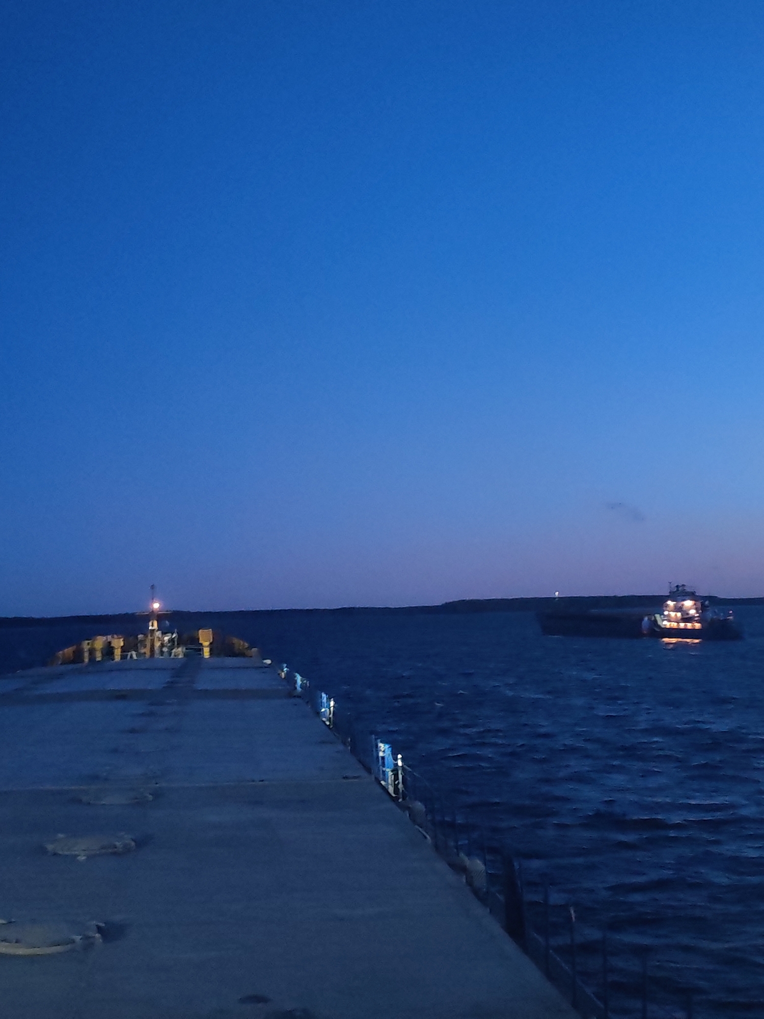 The image shows a pier extending into a body of water, possibly a lake or sea, at dusk or night. A barge or ship with illuminated lights is visible in the distance. The pier appears to be made of concrete, with metal fittings along the edge. The sky is a gradient of blue, indicating twilight or early night. The location is in the city of Unknown, Unknown.
