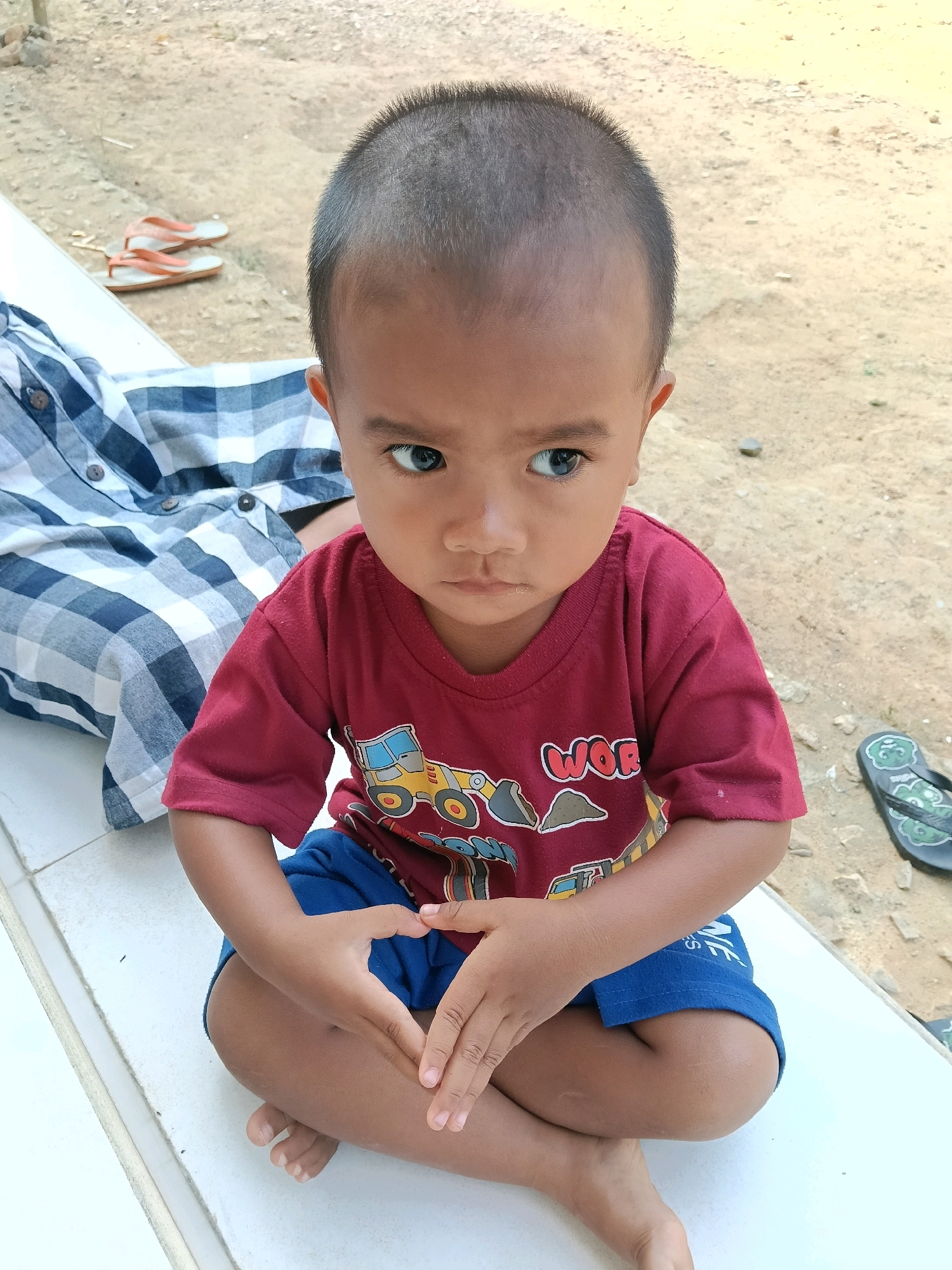 A young child is sitting cross-legged on a white tiled surface. The child is wearing a maroon t-shirt with a graphic of construction vehicles and blue shorts. The child's hands are clasped together on their lap, forming a heart shape. A person wearing a blue and white checkered shirt is seated to the left of the child. The background appears to be an outdoor environment with a dirt ground and some scattered rocks. A pair of orange flip-flops are visible on the ground to the left, and a black flip-flop is on the ground to the right. The lighting suggests it is daytime.