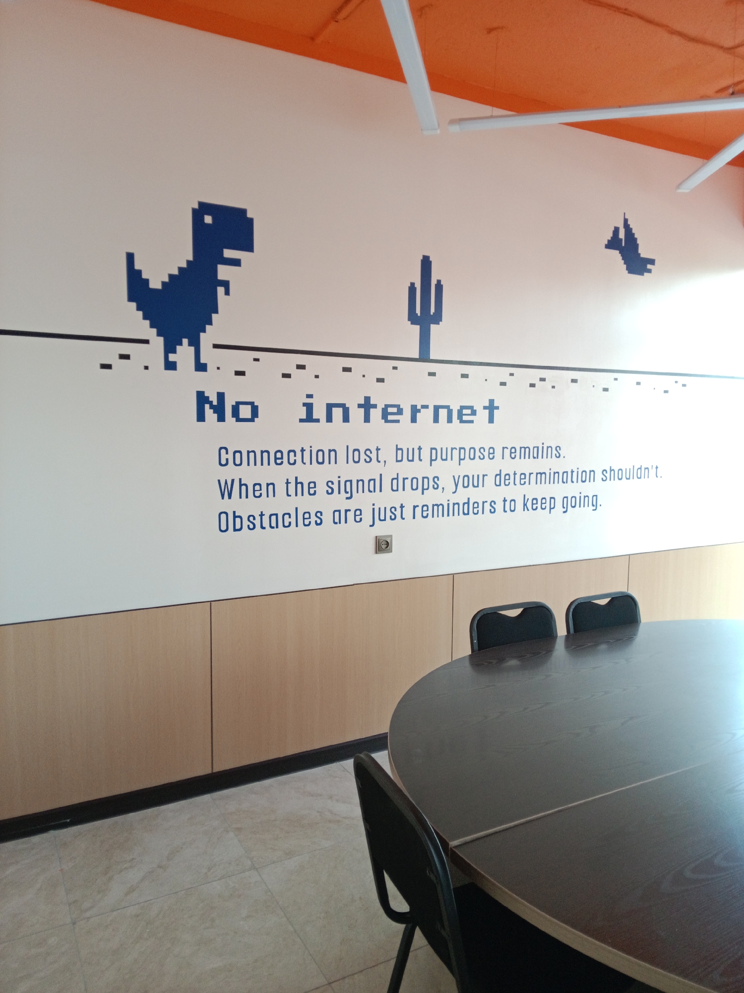 The image shows a meeting room with a mural on the wall. The mural features pixelated graphics of a dinosaur, a cactus, and a bird, along with the text "No internet. Connection lost, but purpose remains. When the signal drops, your determination shouldn't. Obstacles are just reminders to keep going." The room has a large oval conference table with chairs. The walls are painted white above the wooden panels. The ceiling is orange and equipped with fluorescent lights. The floor is tiled. The scene is likely located in an office or co-working space. There are no people visible in the image.
