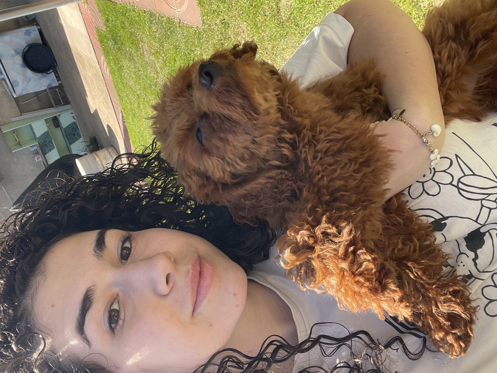 An overhead shot captures a young woman and a reddish-brown, curly-haired dog sharing a peaceful moment outdoors in what appears to be a garden or backyard in Sofia, Bulgaria. The woman is seen from the chest up, her head slightly turned towards the camera, revealing dark, curly hair and brown eyes. She wears a plain white t-shirt, and a delicate gold-toned bracelet adorned with white flower-shaped charms is visible on her right wrist.
The dog, a small to medium-sized breed with dense, reddish-brown curly fur (possibly a poodle mix), rests comfortably on the woman's right arm and chest. Its head is tilted upwards, eyes partially closed, conveying a sense of relaxation or sleepiness.
The setting is bright and sunny, indicated by strong shadows cast across the background. To the right, lush green grass is visible, while to the left, there's a paved area leading to a concrete or stone patio. A white table with a black skillet on it and parts of a building structure are discernible in the distance. The overall impression is one of warmth, affection, and tranquility on a clear, sunny day. There is no visible text in the image.