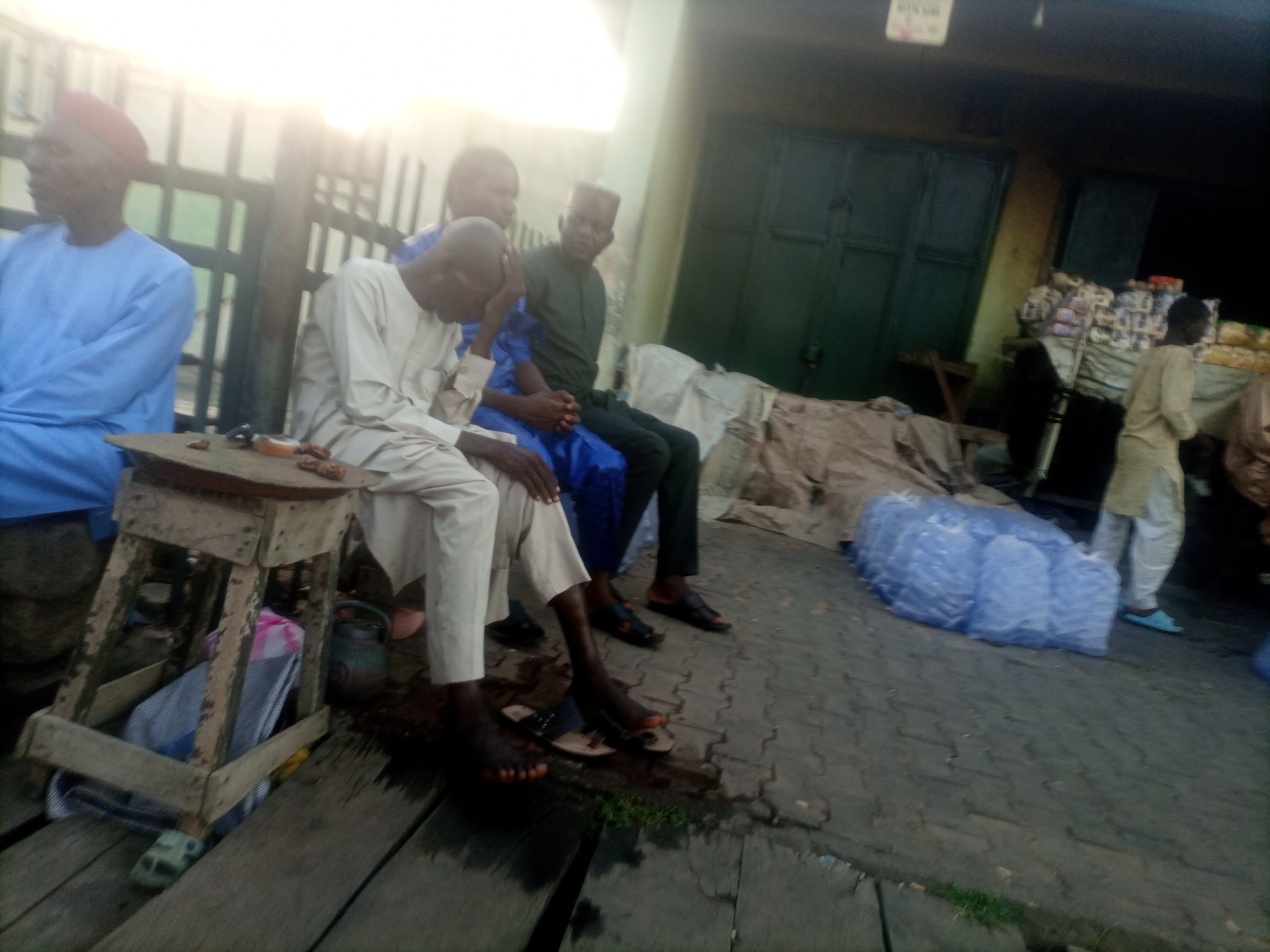 In Arawa, Nigeria, this image captures a group of men gathered outside a shop. The scene appears to be a marketplace, with stalls or shop fronts in the background and what looks like packaged goods on display. The ground is paved with interlocking bricks, and wooden planks form a walkway in the foreground.
Several men are seated on a low wooden structure or bench. One man, wearing a light blue traditional outfit and a red cap, is positioned on the far left. Next to him, a man in a light-colored traditional outfit is seated, facing downwards. Another man in a blue traditional outfit is seated between them. Further to the right, a man in a dark green outfit is also seated. A fifth man, dressed in a light-colored outfit, stands in the background on the right, near some large blue plastic-wrapped items, possibly bundles of goods.
A small wooden table is visible in the foreground, with some items on its surface. The lighting suggests it might be late afternoon or early morning, with a bright light source coming from the left, potentially the sun, casting a glare on the scene. The overall atmosphere appears relaxed, with the men possibly engaged in conversation or resting.