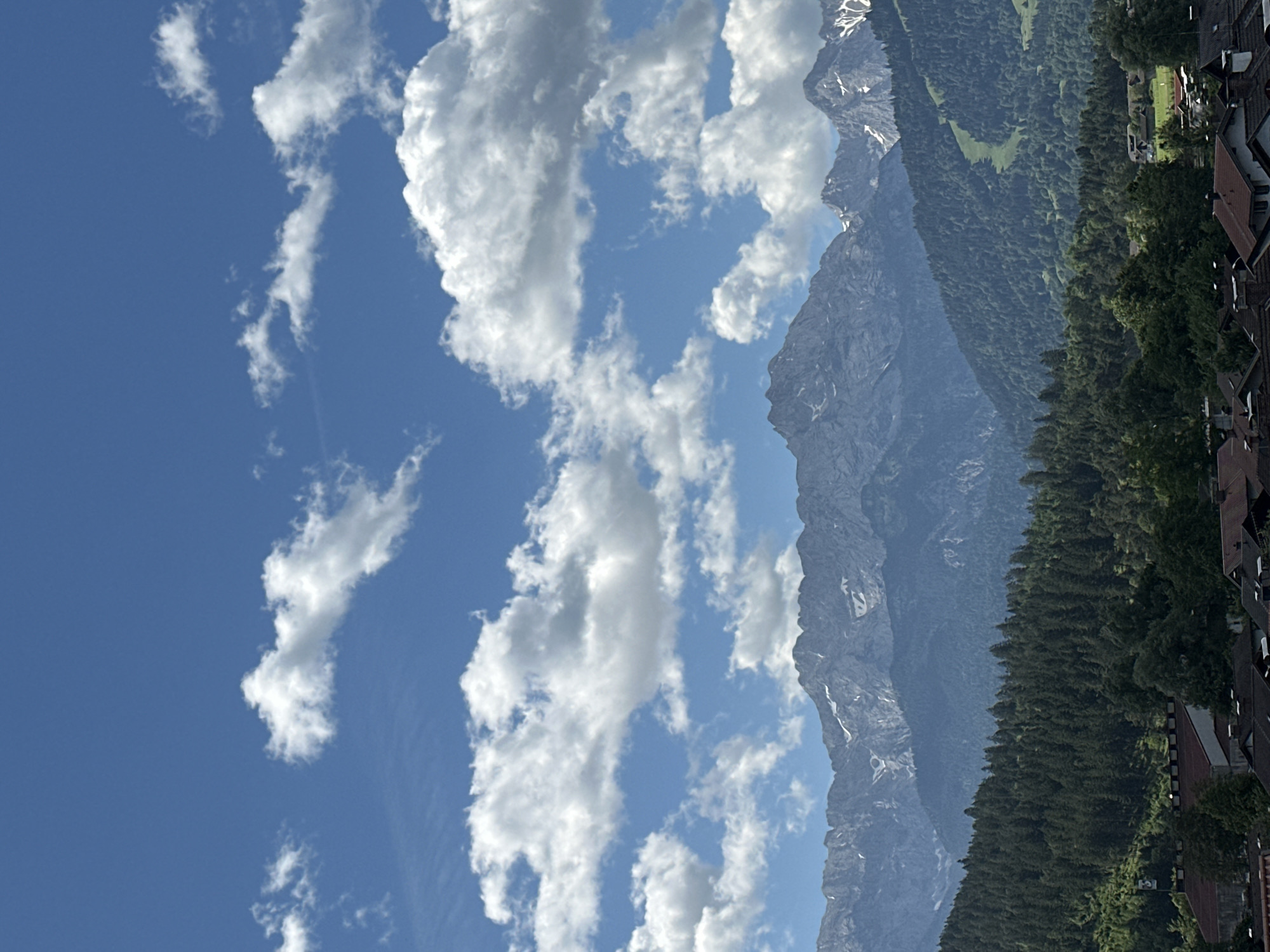 The image depicts a panoramic view of the Zugspitze mountain range, with its jagged peaks partially covered in snow, under a bright blue sky dotted with fluffy white clouds. In the foreground, a dense forest of evergreen trees blankets the foothills, transitioning into a village with red-tiled roofs. A ski slope is visible on one of the mountainsides, suggesting the image was taken during the warmer months, perhaps late spring or early summer. The overall scene evokes a sense of natural beauty and tranquility. The location is the city of Garmisch-Partenkirchen, Germany.