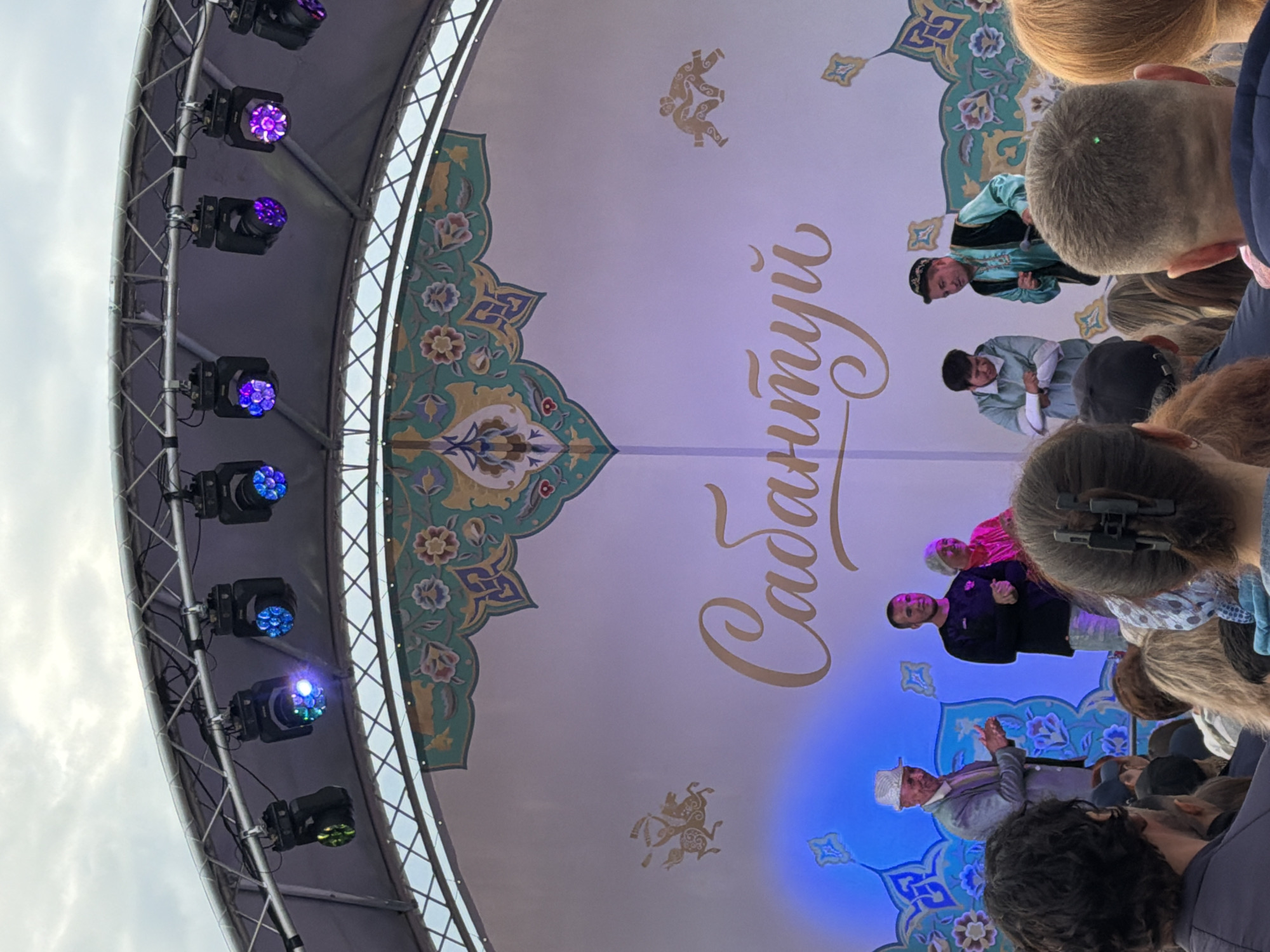 A group of people are on a stage with a white backdrop featuring ornate gold and teal patterns and a large cursive text in gold. In the foreground, a man in a light grey suit and white hat is clapping his hands. To his right, a man in a dark purple shirt and light-colored pants stands with his hands clasped. Next to him, a woman in a bright pink jacket and white head covering is smiling. Further to the right, two men in traditional clothing are on stage. One is wearing a teal vest over a blue shirt and a patterned cap, holding a microphone, and appears to be speaking or performing. The other man, to his left, is wearing a light blue jacket and has his arms crossed. The stage is lit by colorful spotlights. In the background, a crowd of people are visible, watching the performance. The overall setting appears to be an outdoor event.