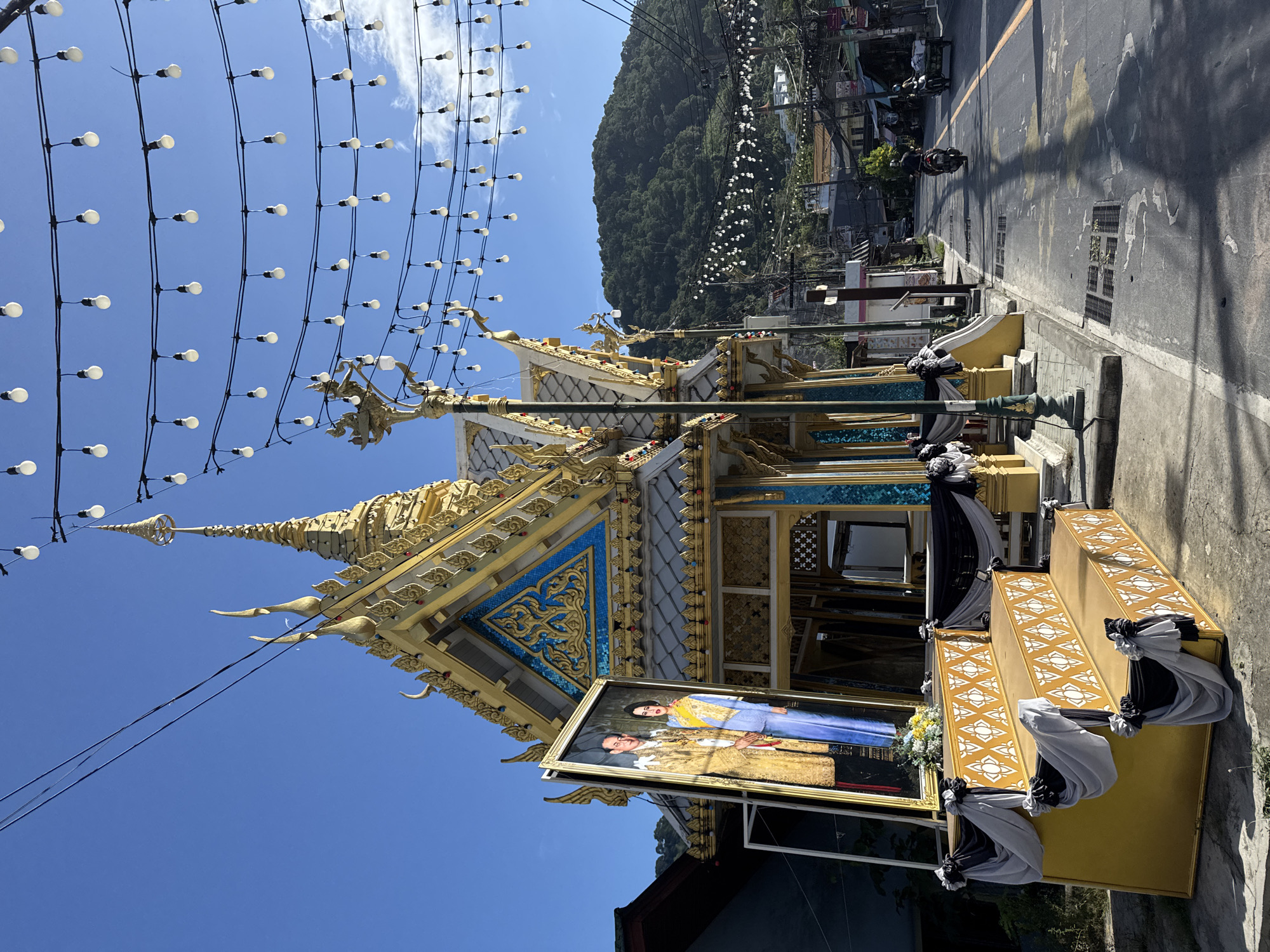 An exterior shot captures a street scene in Patong, Thailand, dominated by a gilded Buddhist structure with ornate tiered roofs and decorative trim. A large framed portrait of a royal couple stands prominently in front of the structure, adorned with black and white fabric drapes. In the foreground, a three-tiered golden platform is also decorated with similar drapes. Strings of white globe lights hang overhead, stretching across the blue sky. The street itself is paved and appears to be a commercial area, with buildings lining one side. Two motorbikes are visible on the road, one with a rider and another pulling a cart. A lush green, tree-covered mountain rises in the background.