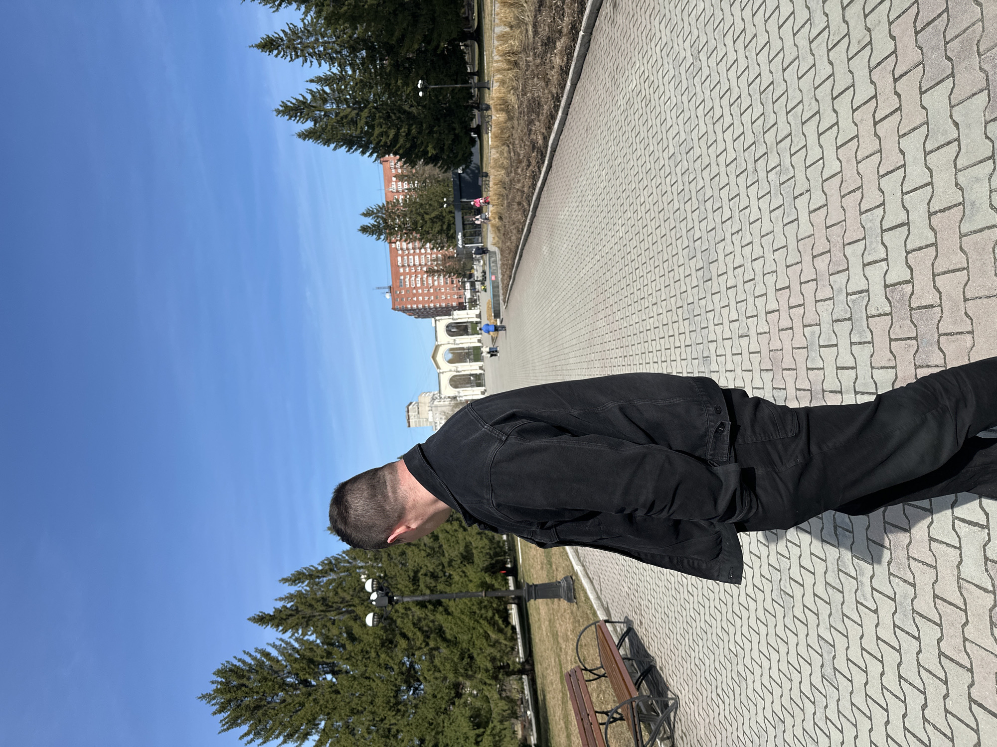 A man stands on a paved walkway in Yekaterinburg, Russia, on a sunny day. He wears a black denim jacket and black pants. The walkway is made of light gray concrete pavers in a repeating pattern. A row of trees lines the walkway. A red brick building and an arched structure are visible in the background. A bench is to the left of the man. The sky is blue.
