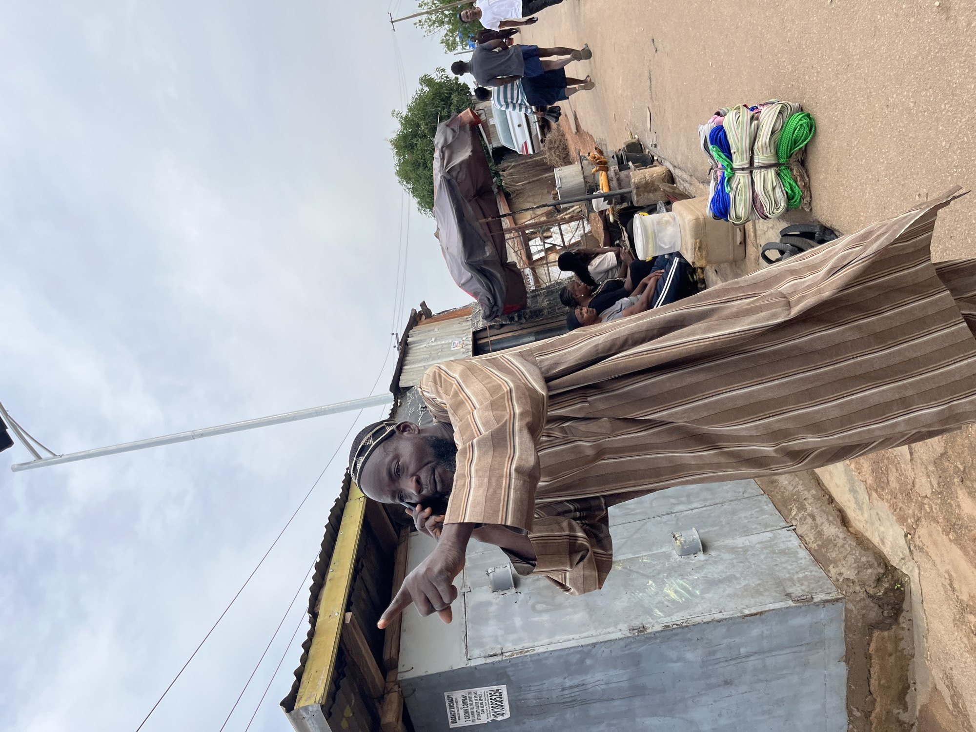 The media file depicts an outdoor scene in Gura Top, Nigeria. The primary subject is a man wearing a brown and white striped garment and a cap, who is on a phone call and pointing with his left hand. In the background, several other people are visible walking on a paved surface and seated near structures. Objects in the scene include bundles of colored ropes, a large white plastic container, various unidentified items covered by a tarp, and what appears to be a building with corrugated metal roofing. The setting suggests a marketplace or a busy street with informal businesses. The lighting indicates it is daytime, with an overcast sky. The overall mood seems casual and everyday. A partially visible sign on a metal structure reads "VACANCY WAKACHI" followed by text that is difficult to fully decipher, but includes "1 CROWN COMPANY", "STUDENTS ABOVE 18 YEARS", and a phone number "28136685254".