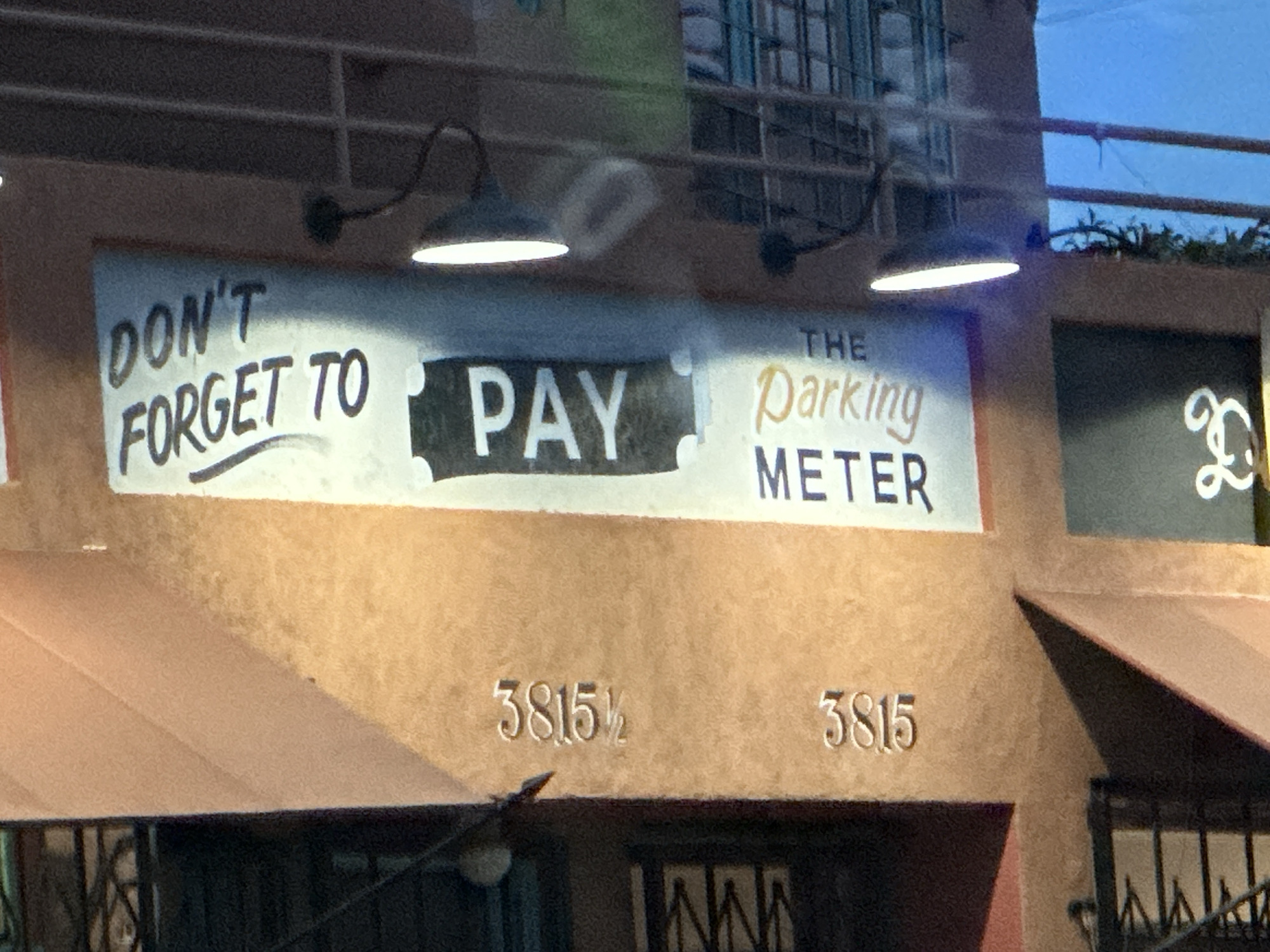 The image depicts a sign attached to a building in what appears to be a commercial district. The sign reads "DON'T FORGET TO PAY THE Parking METER". Below the sign are two addresses etched into the building's facade: "3815 1/2" and "3815". The presence of fractions in the address signifies a place in Los Angeles where many businesses occupy a single property and have their own separate units. The structure of the building suggests a storefront or small business establishment. Two light fixtures are mounted above the sign, illuminating it. The environment appears to be urban, with awnings and building details visible.