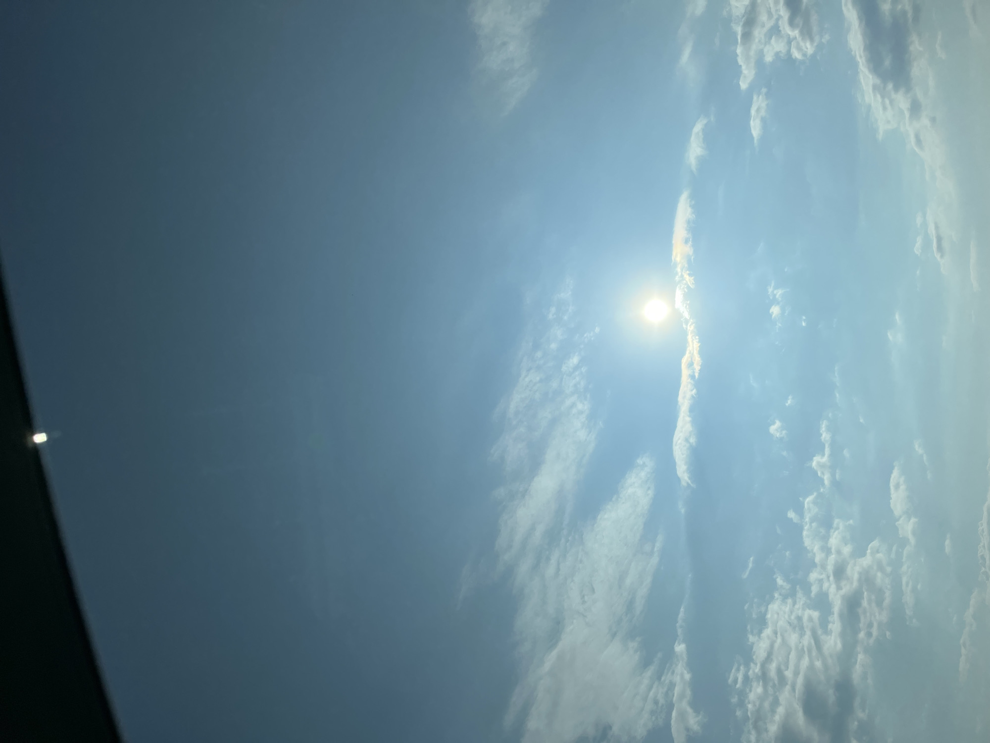 A bright sun shines through a partly cloudy sky, casting light on wispy clouds and a darker, horizontal obstruction at the top of the frame. The image appears to be taken from a moving vehicle, possibly a car, given the obstruction and the upward angle. The sky is a clear blue, with scattered white clouds of varying densities. The sun is positioned slightly to the left of the center of the frame, creating a lens flare effect. The location is not identifiable from the image itself.