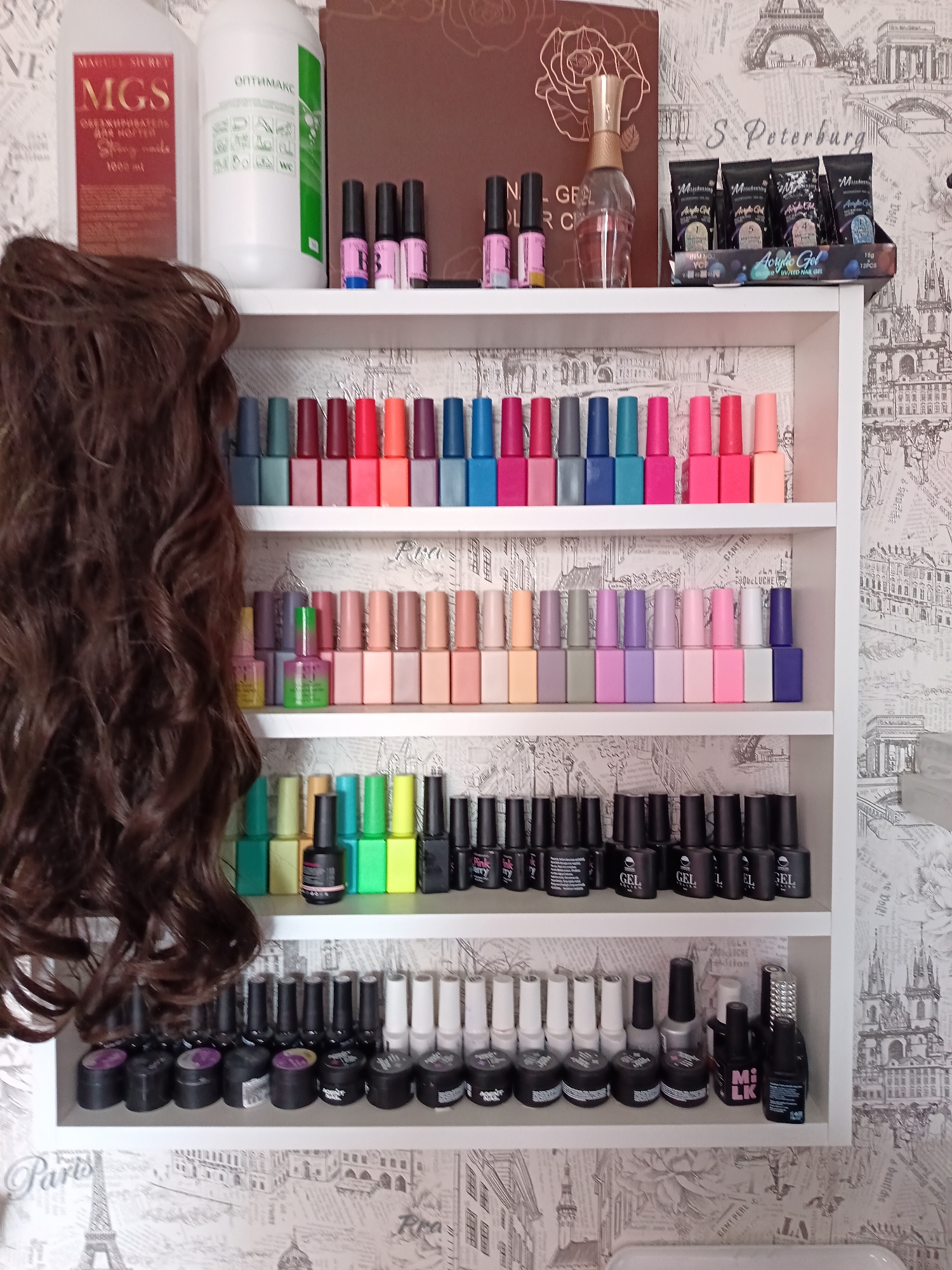 This image presents a detailed view of a personal or professional nail care product collection, neatly arranged on a white, wall-mounted shelving unit. The scene is set indoors against a decorative wallpaper featuring intricate black-and-white line drawings of famous European landmarks like the Eiffel Tower, buildings from St. Petersburg, and Prague, along with city names and other text in English and Russian. The overall location context is Osa, Russia.
On the left side of the frame, a large, dark brown, curly wig or hair extension piece hangs, partially obscuring the shelving unit and its contents.
The top surface of the shelf holds several items. Two large bottles are prominent: one labeled "MAGUL SECRET MGS ОБЕЗЖИРИВАТЕЛЬ ДЛЯ НОГТЕЙ" (Nail Degreaser) for "Strong nails, 1000 ml," and another white bottle marked "ОПТИМАКС" (OPTIMAX) with various usage icons. A brown folder or book with a gold rose design is visible, showing the partial text "GEL." A decorative perfume bottle stands next to it. On the far right, a display stand holds multiple black pouches of "Acrylic Gel UV/LED NAIL GEL," with indications of "15g" and "12PCS." Small pink bottles with a "B" label are also on this top shelf.
The shelves below are densely packed with nail polish and gel bottles, primarily arranged by color. The first shelf features a vibrant array of about 20 bottles in shades of teal, red, hot pink, and orange. The second shelf below showcases approximately 20 bottles in more muted and pastel tones, including beige, light pink, lavender, and soft green. A bright green pot of nail product is also present on this shelf. The third shelf down contains vivid green and yellow polishes, interspersed with black bottles, some labeled "GEL," and partially visible "Pink berry" branding. The bottom shelf is primarily filled with black and white bottles, some clearly marked "MILK," and small black pots of gel, all neatly lined up.