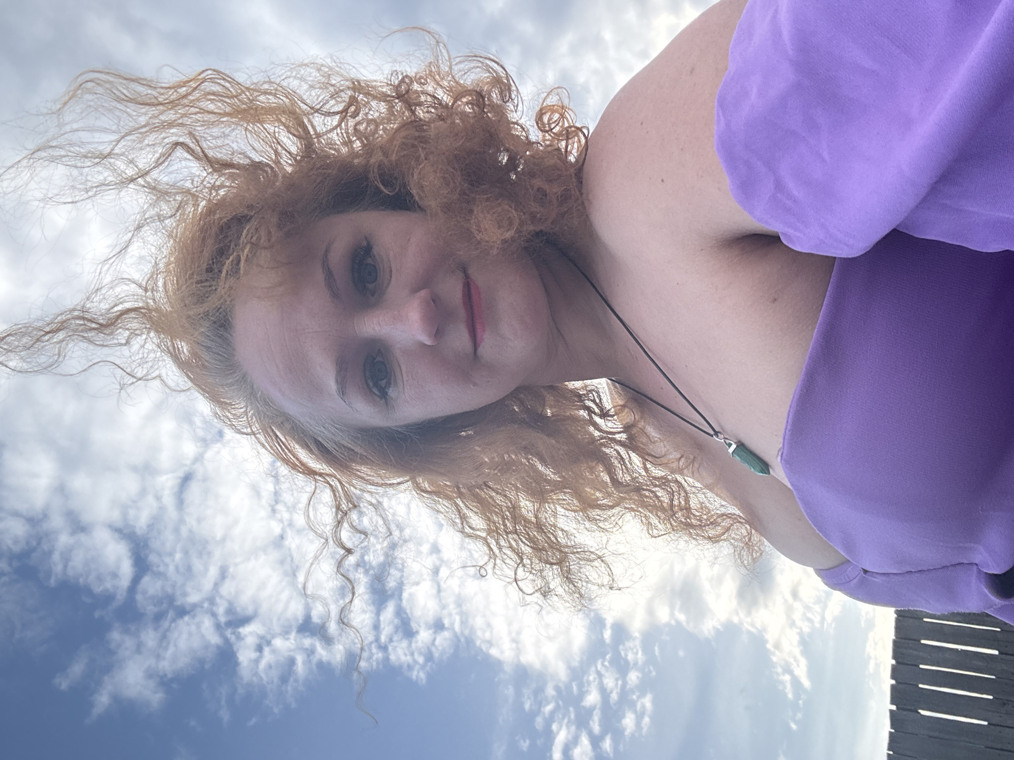 A low-angle outdoor portrait captures a woman with voluminous, windswept curly reddish-brown hair against a bright blue sky dotted with scattered white clouds. The woman, likely in her 30s or 40s, has fair skin, light-colored eyes, and wears pink lipstick. She gazes directly at the camera with a neutral to slightly smiling expression, appearing relaxed.
She is dressed in a vibrant purple garment, possibly an off-the-shoulder top or dress, made of a soft, textured fabric. Around her neck, she wears a necklace featuring a dark cord and a prominent, faceted green gemstone pendant. Her hair is visibly tousled by the wind, suggesting an outdoor environment with a gentle breeze.
The bright daylight indicates it is mid-morning or afternoon, under fair weather conditions. A dark wooden structure, possibly a fence or railing, is partially visible on the far right of the frame, providing a subtle hint of the immediate surroundings. The image conveys a natural, candid moment in an outdoor setting, presumably in Chaikovsky, Russia. No text is visible in the image.