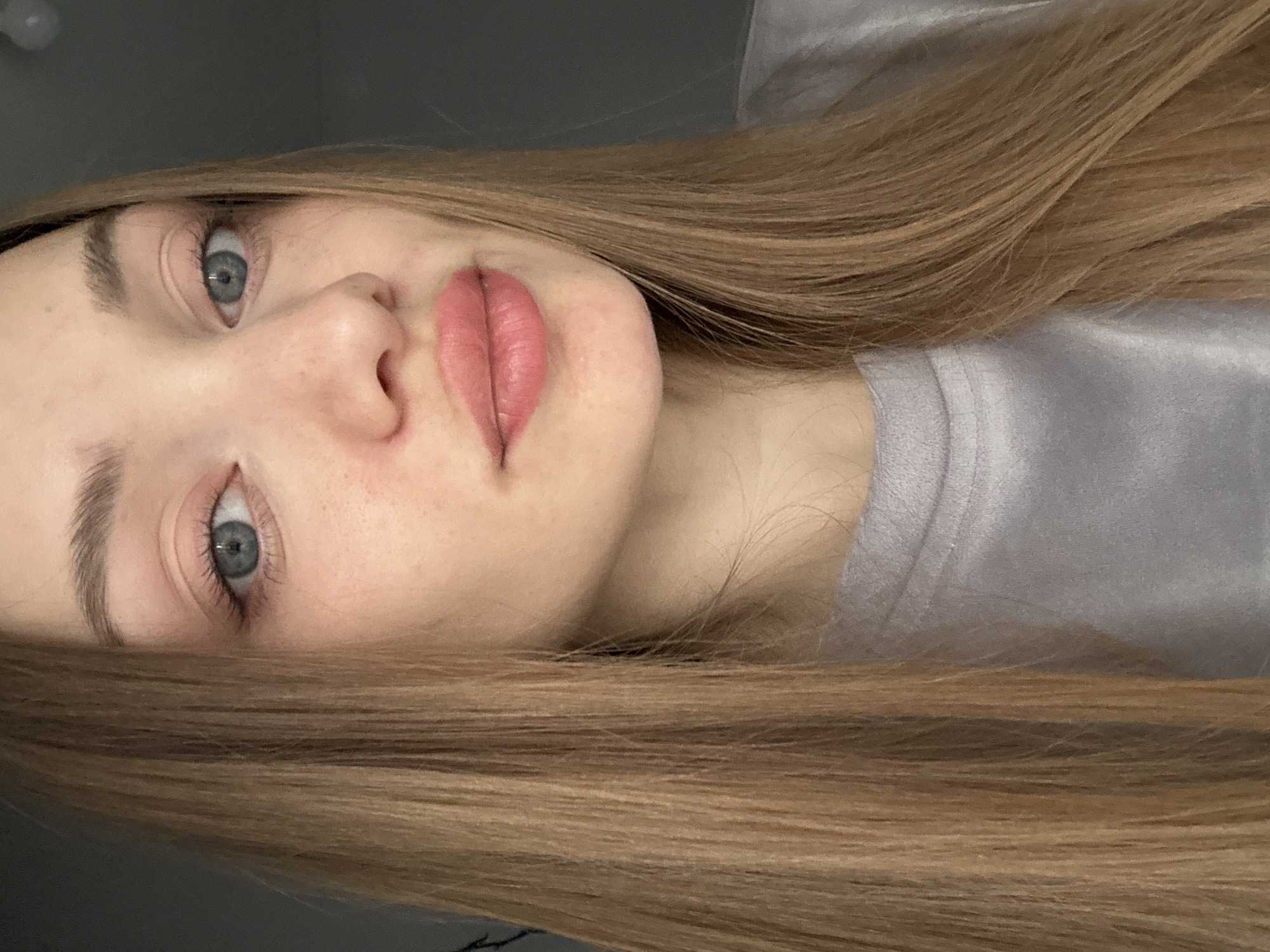 The image is a close-up portrait of a young woman, rotated 90 degrees clockwise, giving the impression that she is lying down with her head positioned towards the left of the frame.
The subject is a woman with fair skin, light blue or grey eyes, and long, straight, light brown hair that cascades around her head and shoulder. Her eyebrows are well-defined. Her lips are full, painted with a pink lipstick or tint, and are slightly parted in a soft, pouting expression. Faint freckles are visible on her nose and cheeks. She is wearing a light grey top, which appears to be made of a soft, possibly satin or velvet, fabric.
Her gaze is directed slightly upwards. The background is dark and out of focus, suggesting an indoor setting with diffused lighting. The overall mood is serene and contemplative, possibly alluring due to her expression. There is no visible text in the image.