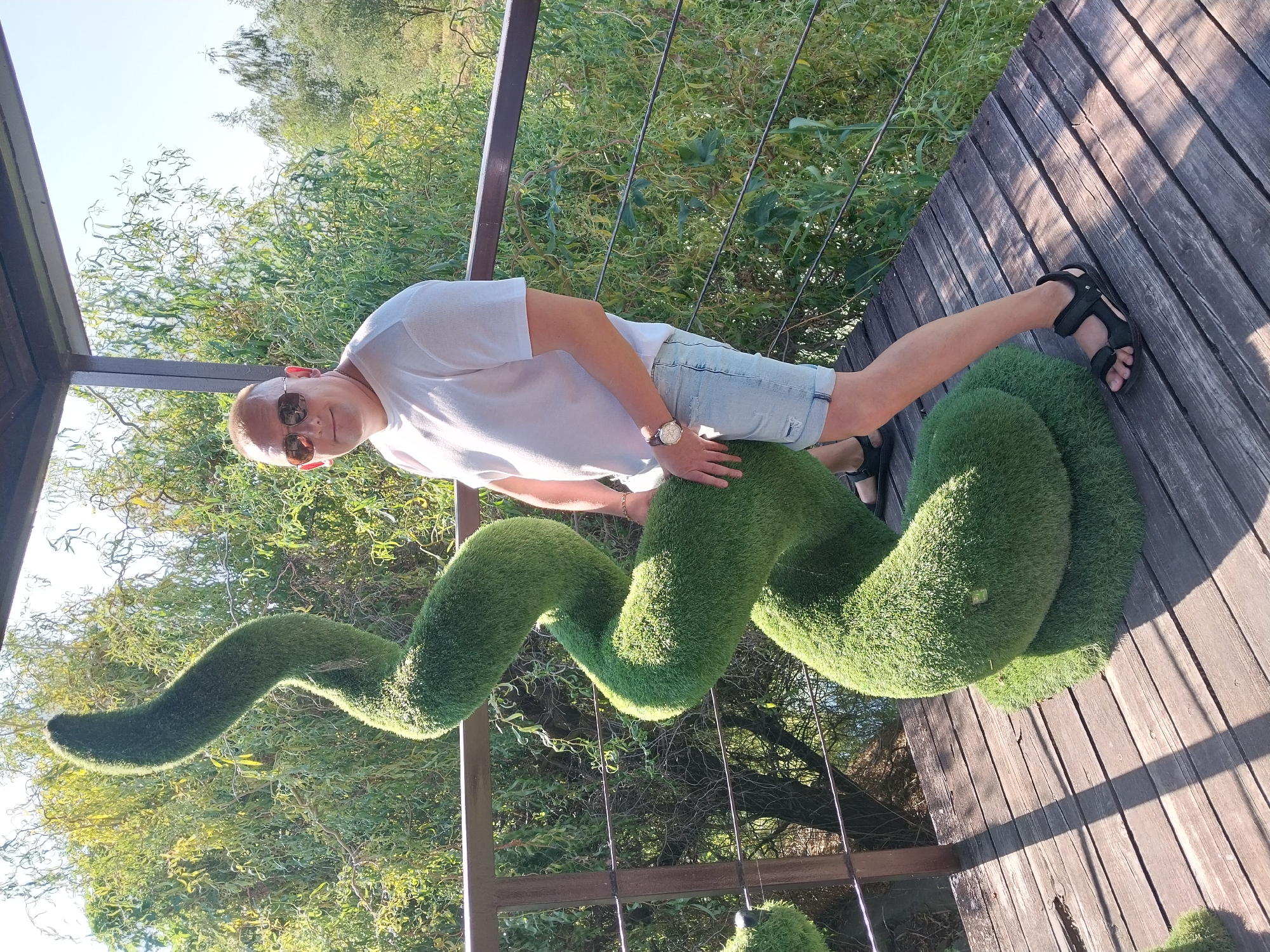 A man wearing sunglasses, a white t-shirt, and denim shorts stands next to a large, spiral-shaped topiary on a wooden deck. The topiary is covered in artificial grass and is several feet tall. The man is smiling and has one hand resting on the topiary. In the background, there is lush green foliage and trees, suggesting an outdoor setting. The wooden deck appears to be part of a balcony or porch, with a railing to the left of the man. The scene suggests a sunny day. The location is Petrykivka, Ukraine.