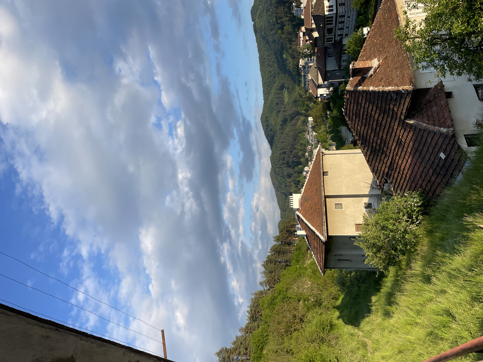 The image presents an elevated perspective of a scene containing residential architecture and natural elements. A small building with a terracotta tiled roof and light-colored facade sits on a grassy incline, partially obscured by greenery. In the background, a densely forested hillside provides a backdrop for buildings that appear to be part of a settlement, possibly a town. The sky is partly cloudy with a mix of blue patches and voluminous grey clouds. Power lines are visible in the foreground. Based on architectural characteristics, the location could be in the city of Zlatograd, Bulgaria. There are no people visible in the scene.