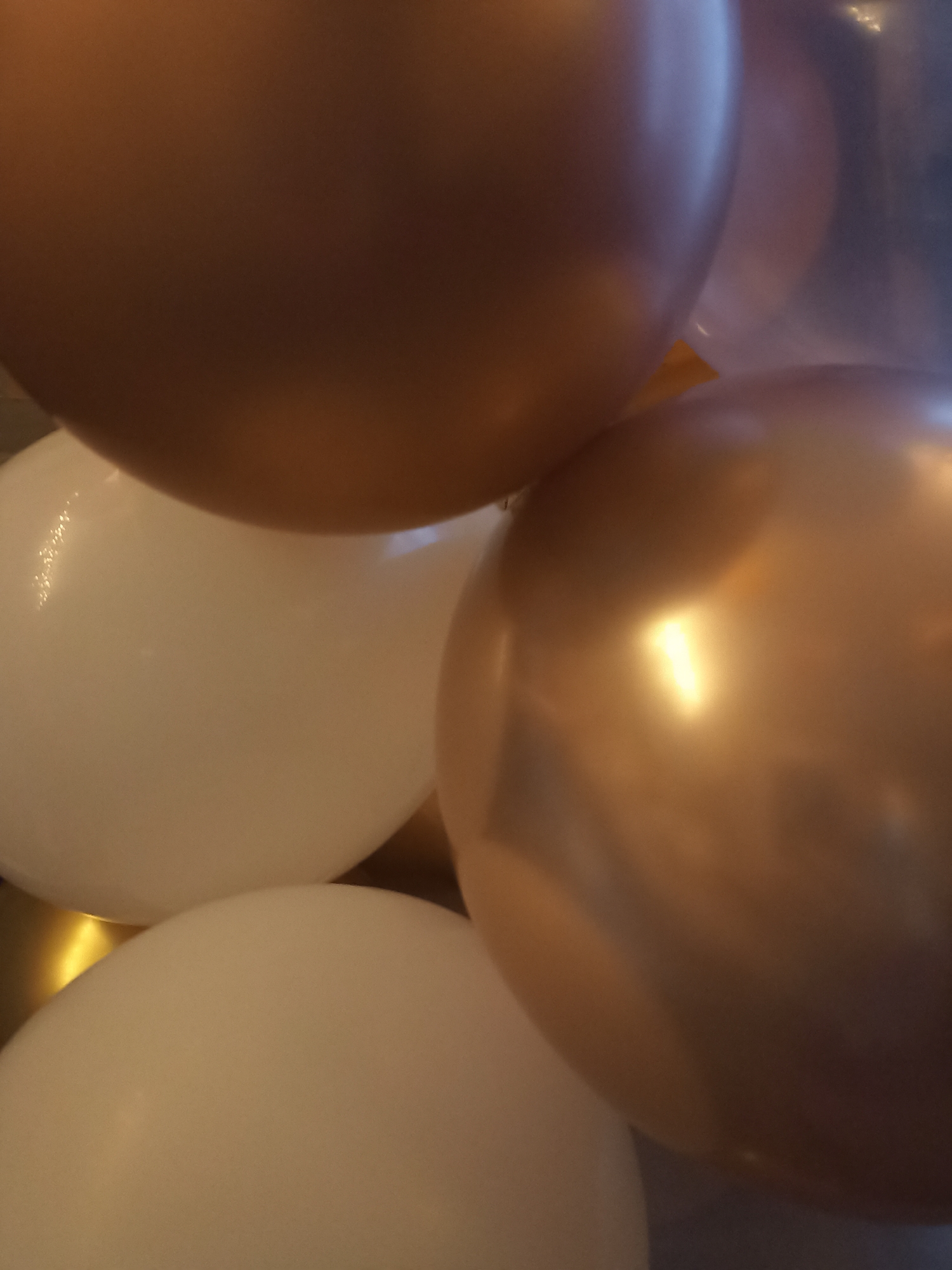 A close-up, indoor, eye-level shot showcases a cluster of balloons. Two dominant balloons are visible: a large, metallic rose gold balloon in the foreground right, and a large, off-white balloon in the lower half of the frame. Partially visible is another off-white balloon to the left of the rose gold one, and a portion of a rose gold balloon at the top center. The balloons are smooth and reflective, with highlights suggesting a light source above and to the right. The background is dark and out of focus, providing no contextual information about the environment or location within Annagry, Ireland. There are no people, actions, or interactions depicted.