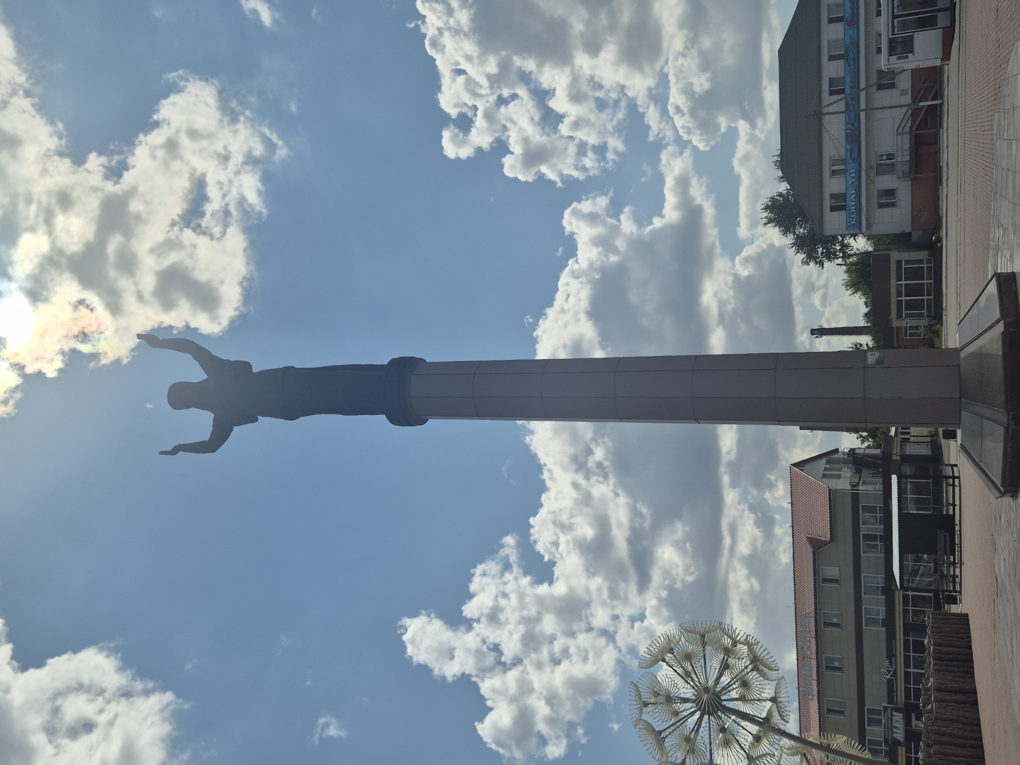 A low-angle, full-shot photograph captures a silhouette of a statue of a person with arms raised against a bright, cloudy sky. The statue stands atop a tall, octagonal pedestal. To the left of the statue, a large, abstract metallic sculpture resembling a dandelion is visible. In the background, a commercial building with a red-tiled roof is situated behind the sculpture, and another building with a blue sign is visible to the right. The scene is set in Kokshetau, Kazakhstan.