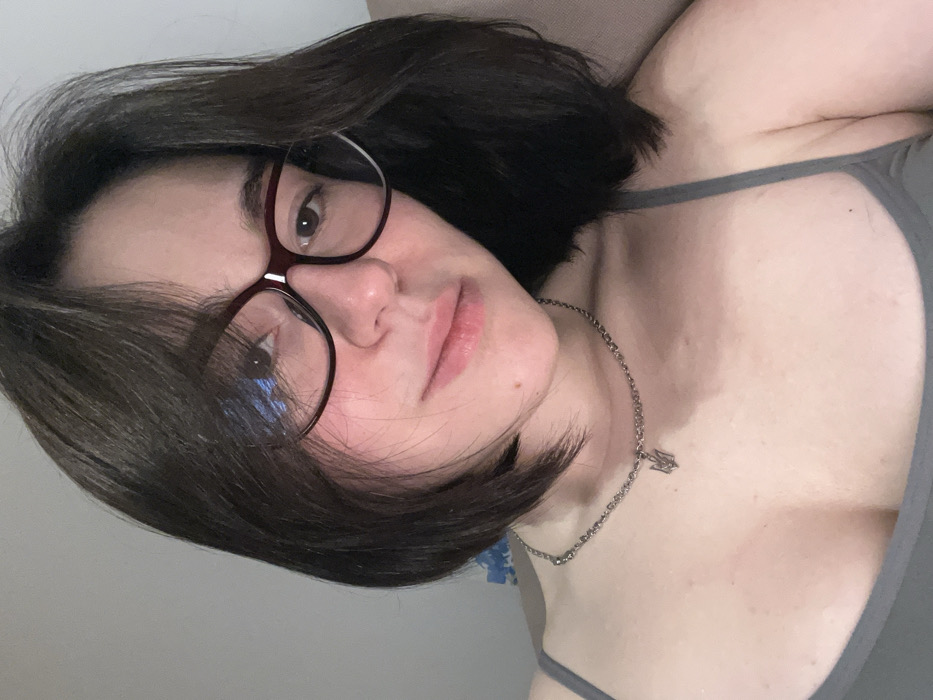 A person with short, dark hair and glasses looks at the camera. They have fair skin and are wearing a gray tank top. A silver necklace with a pendant is visible around their neck. The person appears to be lying down, as the angle is slightly skewed. The background is a plain, light-colored wall.