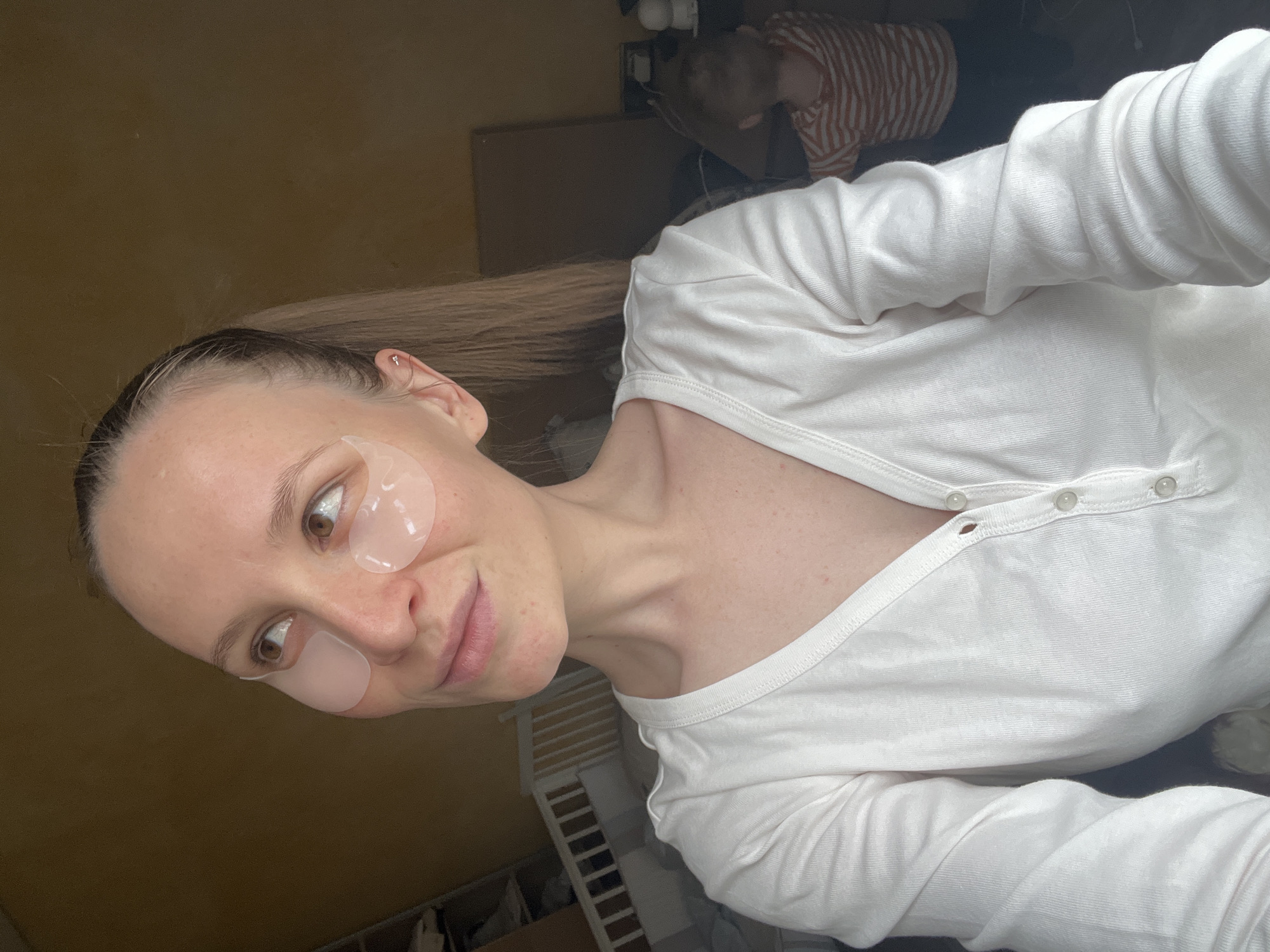 A selfie-style photo features a woman with fair skin and light brown hair. She has under-eye patches applied. The woman is wearing a white, button-down, long-sleeved shirt. In the background, a second person is partially visible, wearing a striped shirt. Part of a white shelving unit is also visible. The location of the photo is reported as the city of Khimki, Russia.