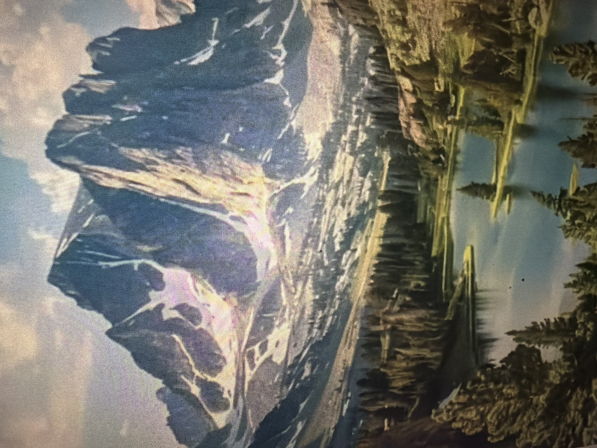 The image depicts a majestic mountain landscape with a serene lake in the foreground. Towering, jagged peaks, partially covered in snow and illuminated by warm sunlight, dominate the background. The slopes leading down from the mountains are a mix of rocky terrain and lush green vegetation. A tranquil lake, reflecting the sky and surrounding trees, occupies the mid-ground, with small, wooded peninsulas extending into its waters. A dense forest of evergreen trees lines the shores of the lake, leading the eye towards the imposing mountain range. There are no people or animals visible in the image. The scene evokes a sense of natural grandeur and peaceful isolation. This image is not located in Baan Mae Nam, Thailand.