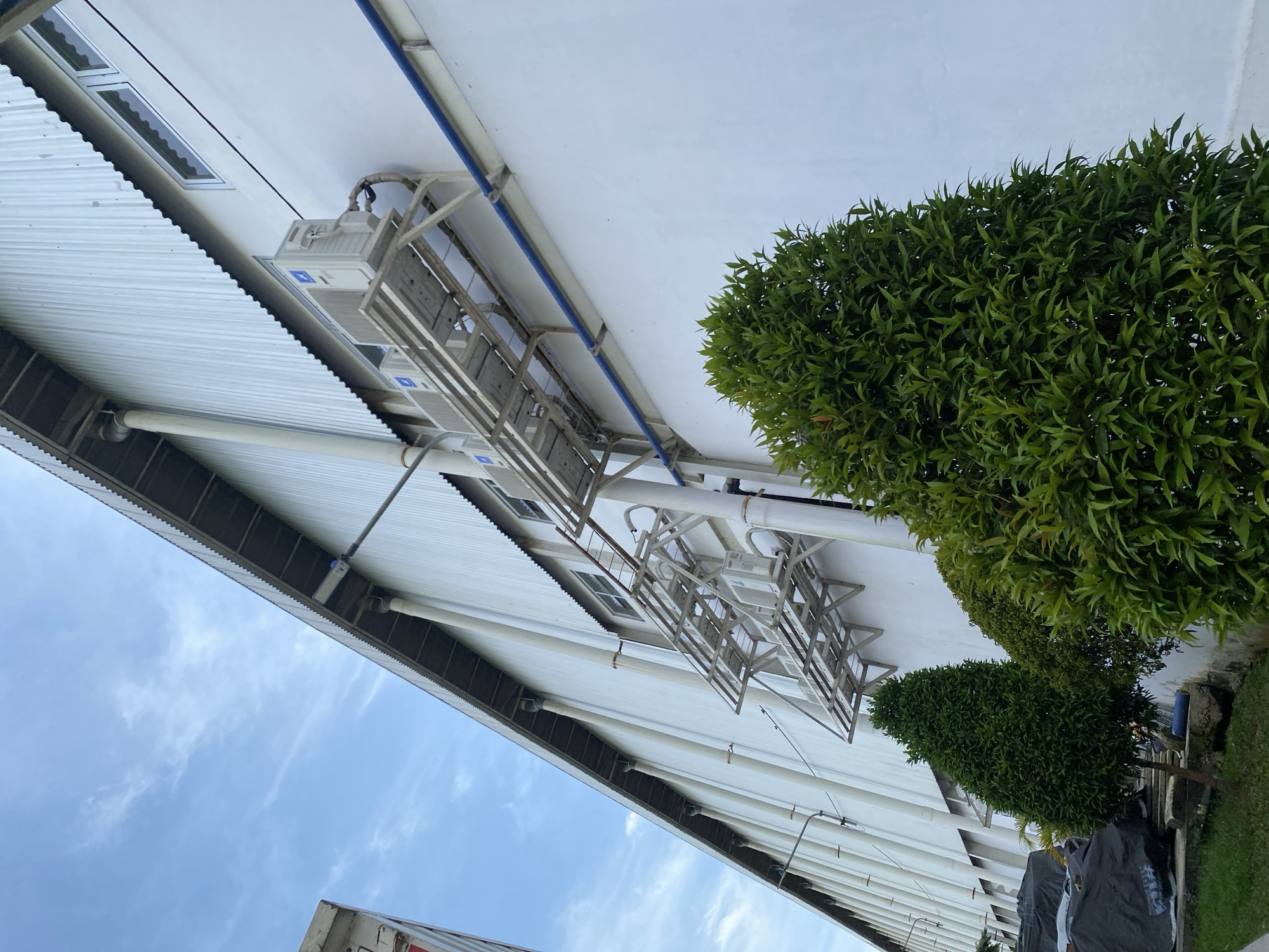 The image shows the exterior of a large, white building with a corrugated metal roof in Gambut Barat, Indonesia. The building has multiple windows and several outdoor air conditioning units mounted on metal racks. A blue pipe runs horizontally along the wall. In the foreground, there are two well-maintained green bushes. The sky above is blue with some wispy clouds. The overall scene depicts the side of an industrial or commercial building.