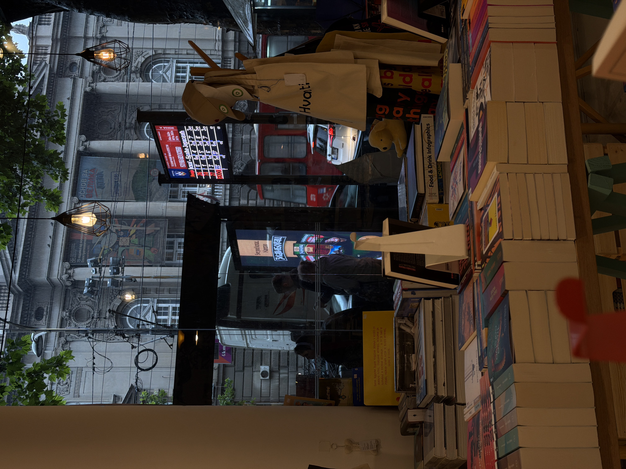 This image captures a street scene in Vračar Urban Municipality, Serbia, viewed through the window of what appears to be a bookstore or shop. The foreground is dominated by stacks of books displayed on wooden tables, with their spines facing outwards. Among the books, a yellow plush toy dog is visible, placed on a stack of books titled "Food & Drink Infographics".
Through the window, the exterior environment is seen, featuring a grand, old building with classical architecture, including columns and ornate detailing. There are large posters on the building's facade. A digital display board shows a grid of numbers and text in Cyrillic script, possibly a schedule or score. Overhead, two industrial-style pendant lights hang, casting a warm glow.
Several people are visible through the glass, seemingly in transit or within another establishment. A white van and a red bus are parked or moving on the street. The atmosphere appears somewhat overcast, with a hint of rain or moisture. Bags, including tote bags with the word "Hvati" printed on them, are displayed hanging on a rack. The overall impression is a blend of urban street life and a cozy, book-filled interior.