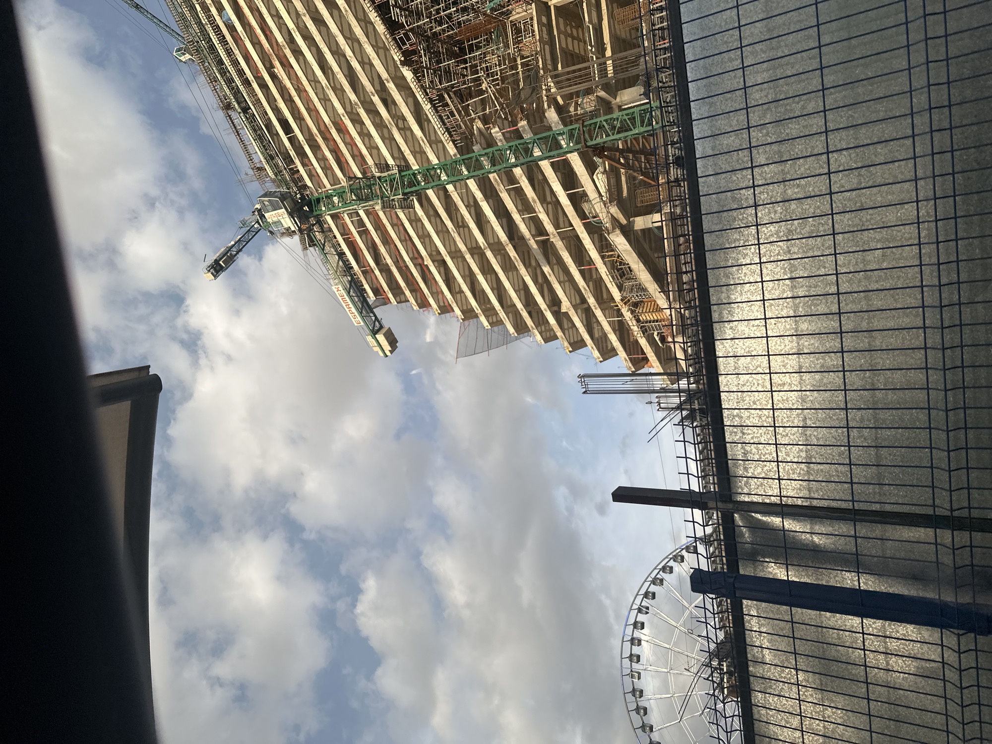 A construction site for a tall building is shown, with a Ferris wheel in the background, indicating the location is likely near a recreational area or entertainment venue in Puebla, Mexico. The building under construction is partially completed, with visible concrete structures, rebar, and scaffolding. A green construction crane is prominent, extending over the building. The scene is viewed from behind a metal fence, suggesting the perspective is from a public area or a neighboring property. The sky is partly cloudy, with sunlight illuminating the construction site. There are no people clearly visible in the frame.