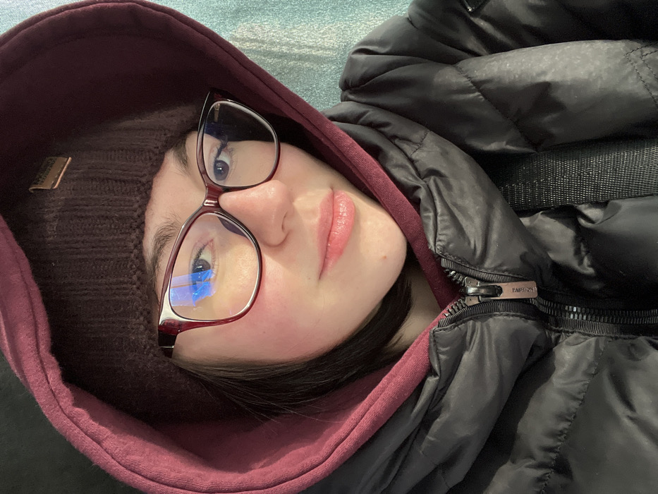 A young woman is depicted in a close-up, chest-up selfie format, centered within the frame. She wears dark-rimmed glasses, a dark brown knitted beanie with a small rectangular tan label, a burgundy hooded sweatshirt with the hood pulled up, and a black puffer jacket, partially zipped, with the zipper pull featuring the word "FASHION." Her dark hair is visible around her face, and her expression is neutral with a slight smile. Reflections of blue light are visible on the lenses of her glasses. A dark strap, possibly from a bag, is visible over her right shoulder. The background is an out-of-focus, textured, light teal or blue surface. The scene takes place in Uman, Ukraine.