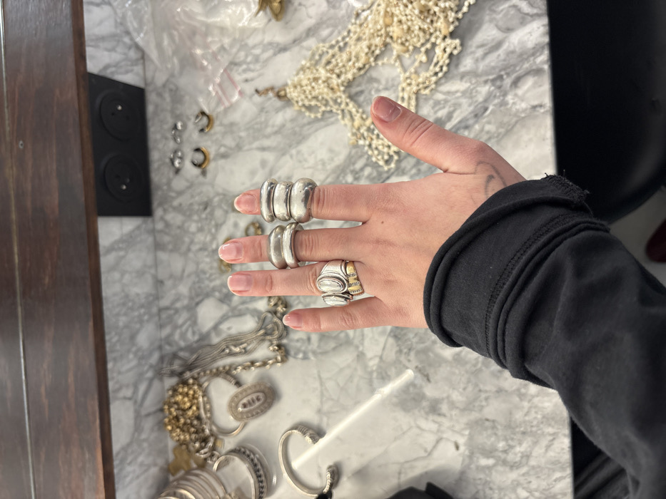 A hand wears three substantial rings: one on the index finger, another on the middle finger, and an elaborate ring on the ring finger, a combination of silver and gold elements. Below are assorted pieces of jewelry including necklaces, earrings, and bracelets, scattered on a marble surface. A section of a wooden dresser and a portion of a black article of clothing are also visible.