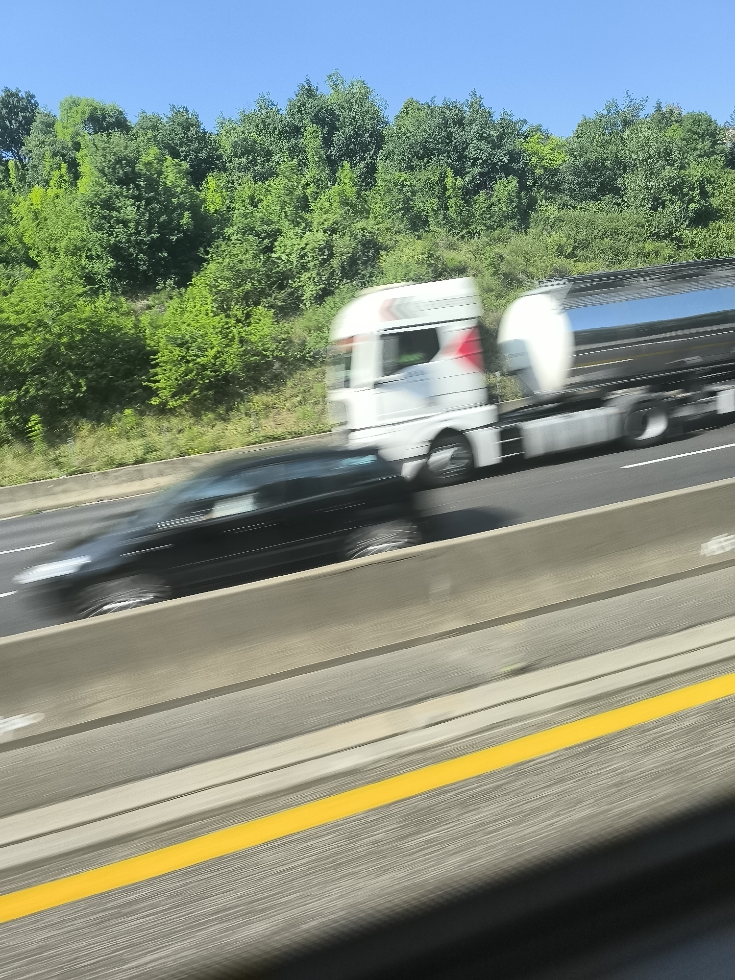 The image shows a highway scene in L'Horme, France, on a sunny day. A white semi-truck with a tanker trailer is traveling on the road. A black car is moving in the adjacent lane, appearing blurred due to motion. Green trees and vegetation line the background. A yellow lane marker is visible on the road surface. The image seems to have been taken from a moving vehicle.