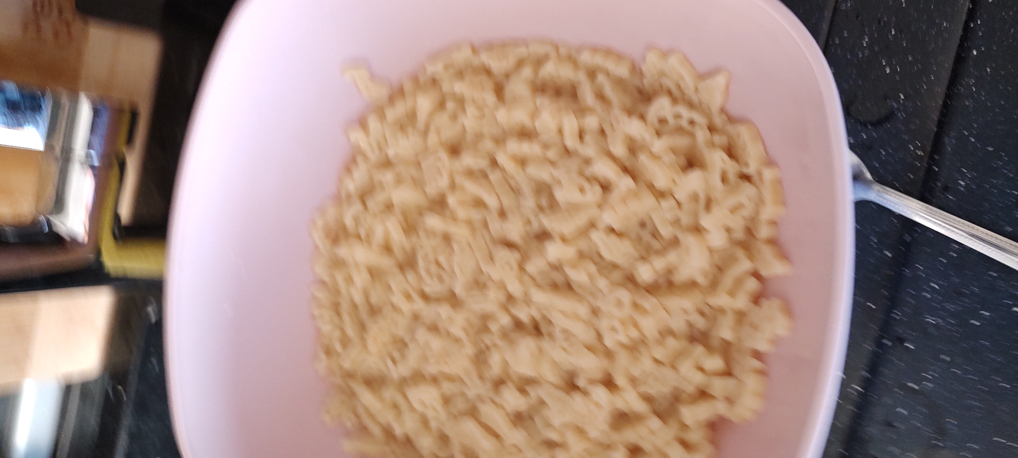 A blurred close-up shows a pink bowl filled with alphabet pasta. To the left, a portion of a counter surface is visible, along with what appears to be a reflective object. A silver spoon rests against the right edge of the bowl on a dark countertop.