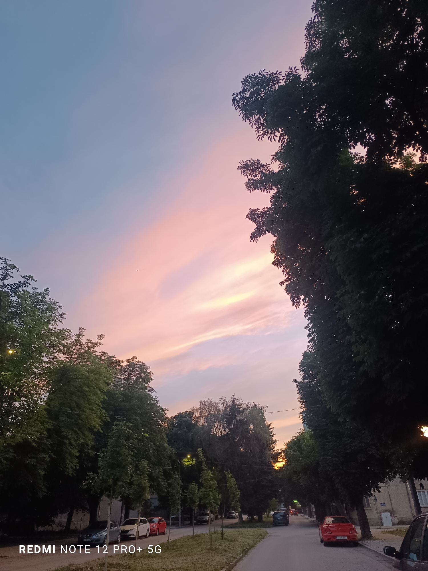 This media file depicts a street scene in Targovishte, Bulgaria, during a sunset. The sky is filled with soft hues of pink and blue, indicating the transition from day to night. Tall trees line both sides of a paved road, their dense foliage creating a canopy effect. Several cars are parked along the street and on the side of the road, including a red car prominently featured in the foreground and a few other vehicles further down the street. Buildings, partially visible behind the trees, suggest a residential or urban setting. The lighting is dim, characteristic of dusk, with a few streetlights visible and the warm glow of the setting sun in the distance. The overall mood is peaceful and serene, capturing a quiet moment in the city as day ends. The text "REDMI NOTE 12 PRO+ 5G" is visible in the bottom left corner of the image, indicating the device used to capture the photograph.