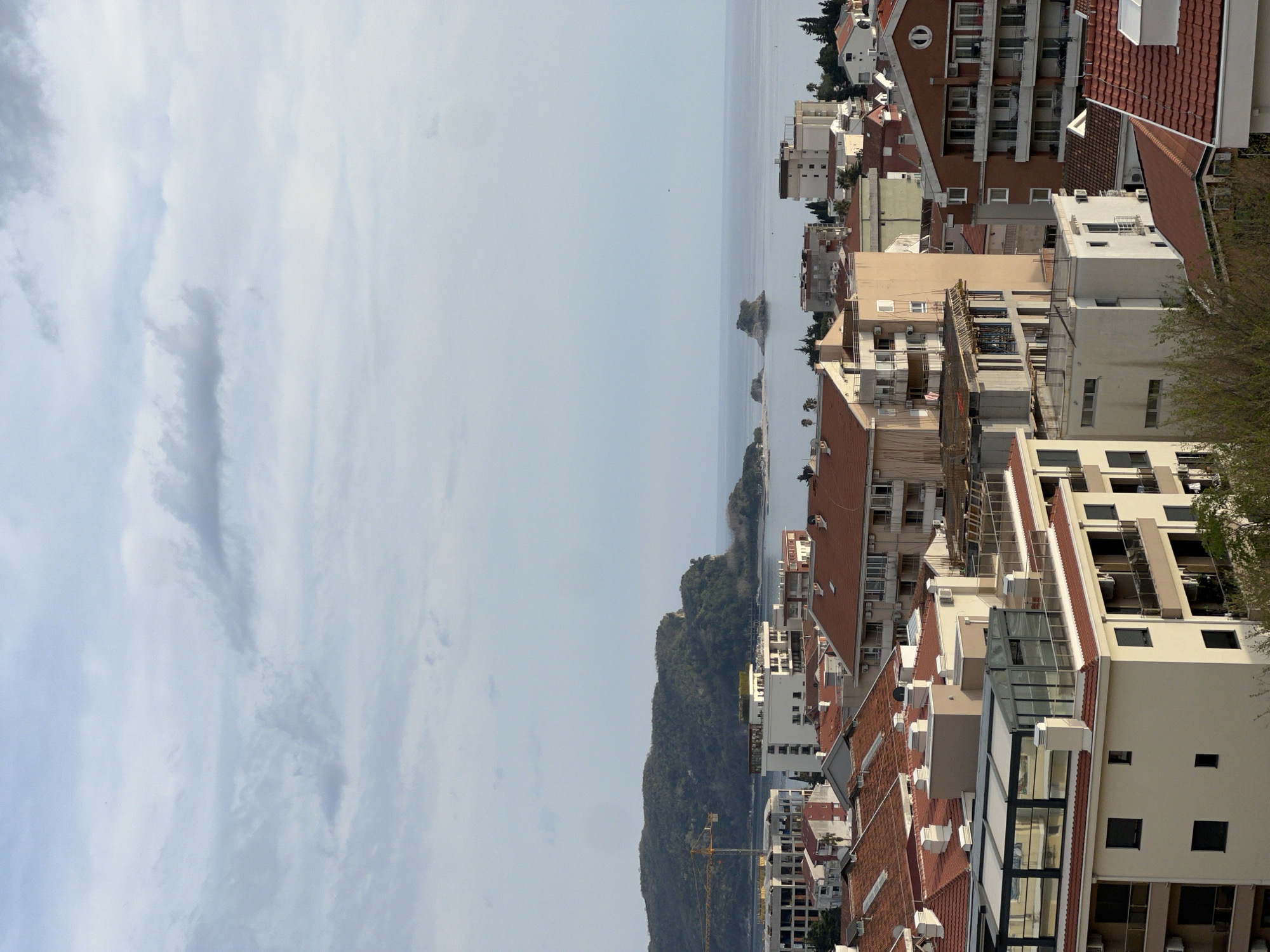 This is an aerial view of the city of Budva, Montenegro. The image is oriented with the sky and clouds on the left side and the city buildings and the Adriatic Sea on the right. The buildings are densely packed, with varied architectural styles. Red tile roofs are prominent among the buildings. In the distance, there are small islands along the coast. A forested hill is also visible. A construction crane is visible near some buildings. The Adriatic Sea is relatively calm. The sky is overcast.