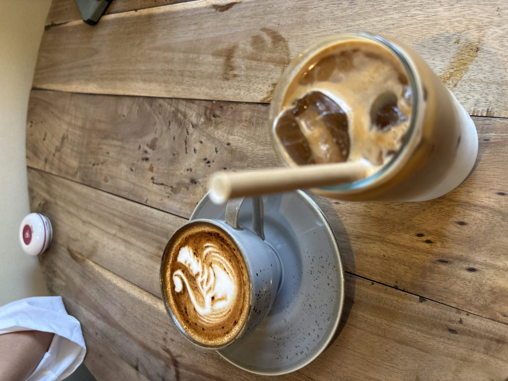 The image shows two drinks on a wooden table, seemingly at a cafe in Kuta Selatan, Indonesia. On the left is a cappuccino with latte art in a light grey cup and saucer. A paper straw rests on the saucer, partially obscured by the cup. To the right of the cappuccino is a glass of iced coffee, also with a straw. In the background, there is a small white call button adhered to the wall and what appears to be an edge of a white fabric. The wooden table's surface has visible grains and a worn look. The scene suggests a coffee break.