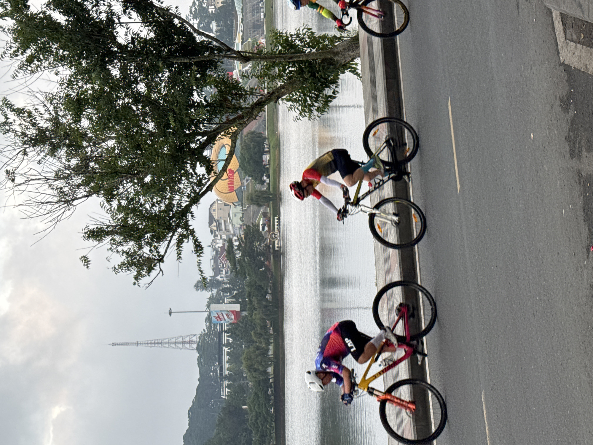 The image depicts a cycling race on a paved road adjacent to a body of water, likely Xuan Huong Lake in Phường Xuân Hương, Đà Lạt, Vietnam. In the foreground, three cyclists are actively participating in the race. The cyclist on the left wears a white helmet, a multi-colored cycling jersey with "LAP" visible on the back, black shorts, and white socks. This cyclist is on a bicycle with a yellow frame and orange accents on the wheels. The second cyclist from the left wears a red and white helmet, a yellow and red cycling jersey, black shorts, and lime green socks. This cyclist is on a bicycle with a yellow frame and black wheels. The third cyclist, partially visible on the right, wears a blue and green cycling jersey and a blue helmet. In the background, across the water, the cityscape of Đà Lạt is visible. This includes a prominent tower, trees, buildings with red roofs, and a distinctive, large, golden dome-shaped structure. The sky is overcast and gray, suggesting a cloudy day.