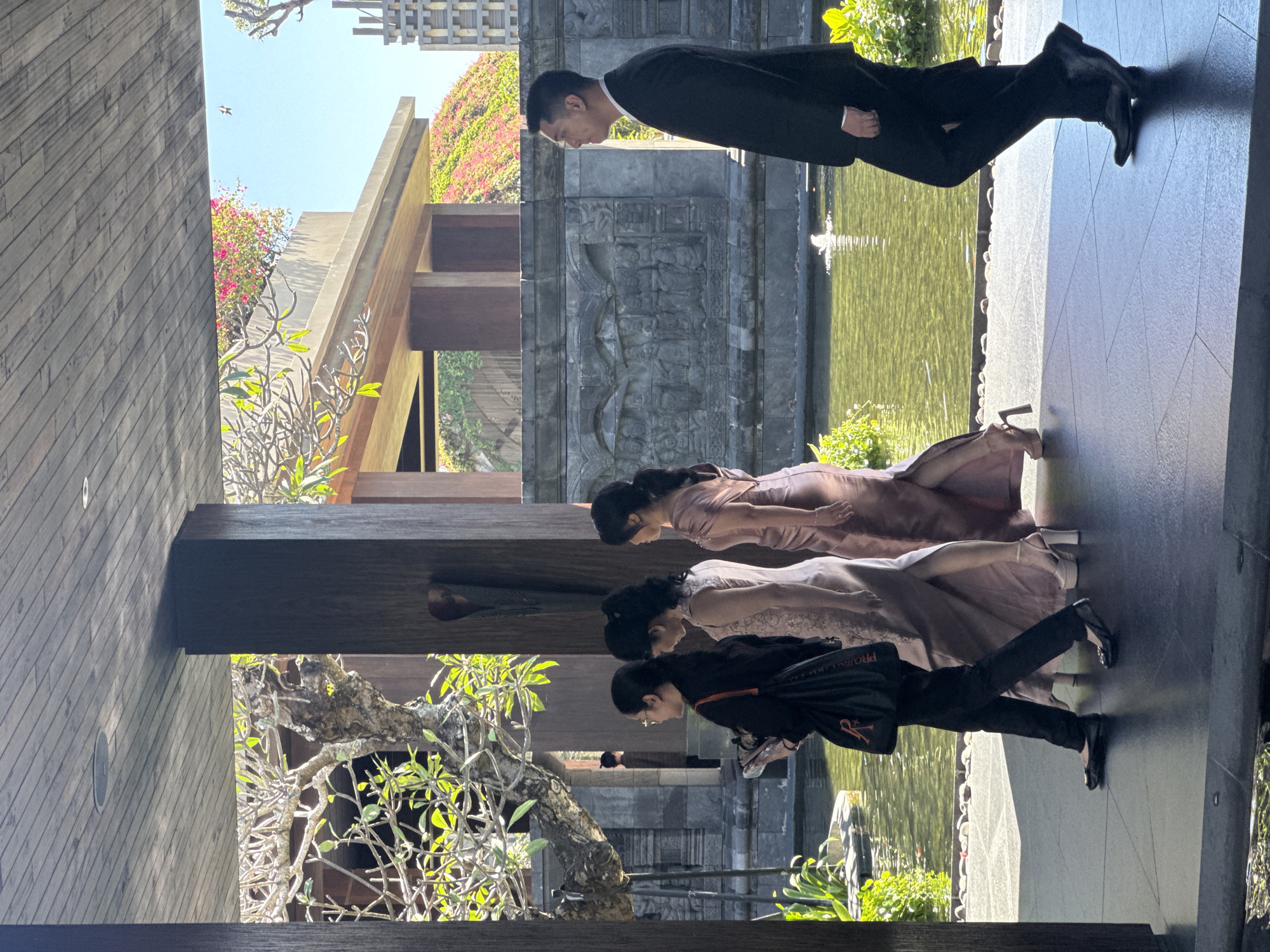 Captured in Kuta Selatan, Indonesia, the image presents four individuals near a building adjacent to a body of water. The first person, a male wearing a dark suit, stands closest to the water's edge. Behind him, two women are seen in rose-toned dresses. Further back, a fourth person, also dressed in dark attire and carrying a black bag with an orange emblem, stands nearest to a tree. The building features a carved stone facade, wooden support beams, and a section with green vegetation. The environment includes both cultivated and natural elements.