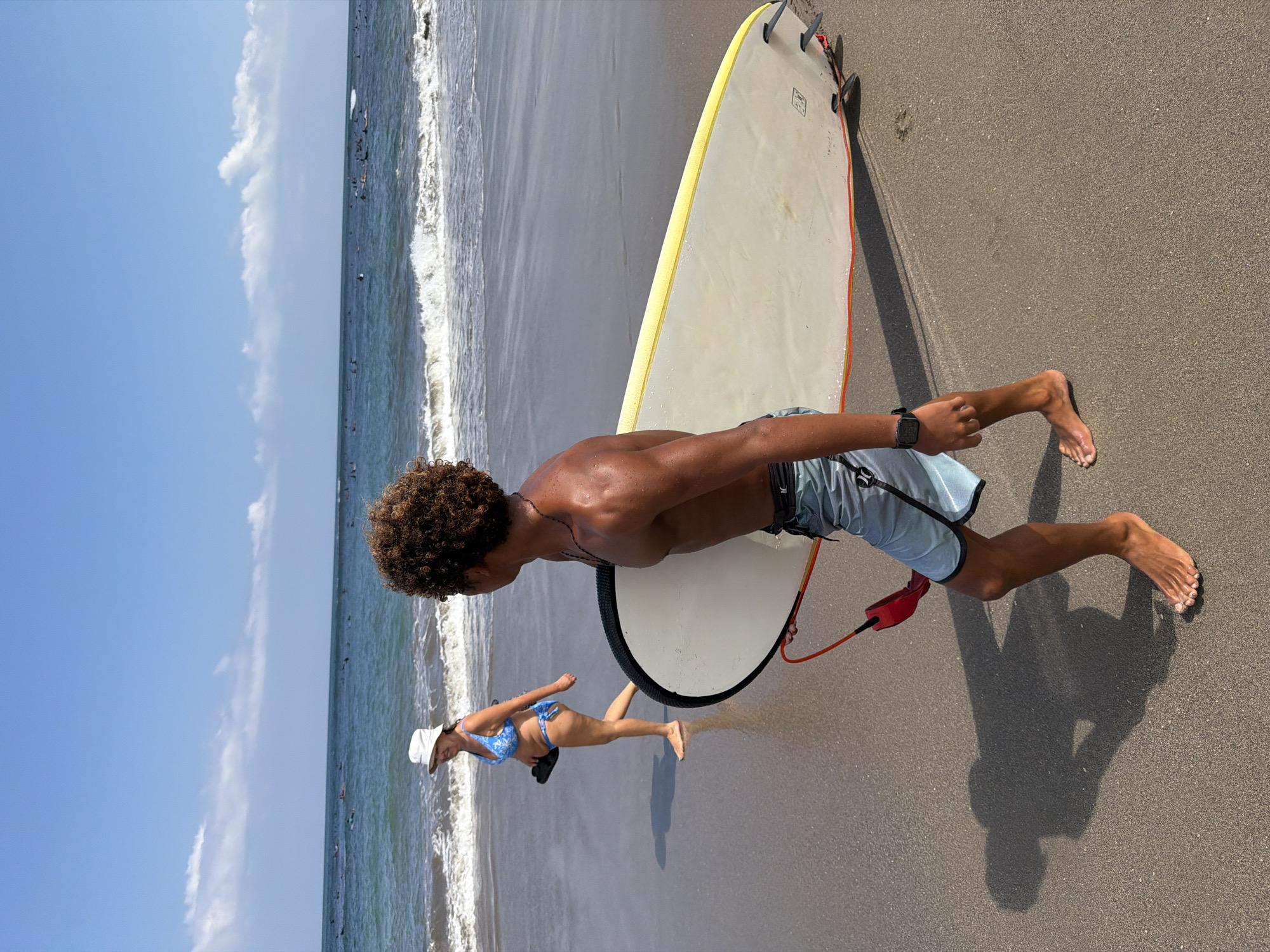 In North Kuta, Indonesia, two individuals are depicted on a sandy beach under a clear blue sky with scattered white clouds. A person in the foreground, viewed from the back, is a young male with curly dark hair and tanned skin. He is shirtless and wearing light blue patterned swim shorts. He is carrying a white surfboard with a yellow edge and a red leash. His left arm is bent, holding the leash, and a black smartwatch is visible on his wrist. His shadow stretches out before him on the sand.
Further back, walking towards the left of the frame, is an older female wearing a white hat and a blue and white patterned bikini. She is barefoot and carrying a pair of dark flip-flops in her left hand. Her body is angled away from the camera as she walks along the wet sand near the shoreline.
The ocean stretches out beyond the beach, with gentle waves breaking on the shore. Numerous surfers are scattered across the water, some riding waves and others waiting for them. The overall scene captures a typical day at the beach with recreational activities underway.