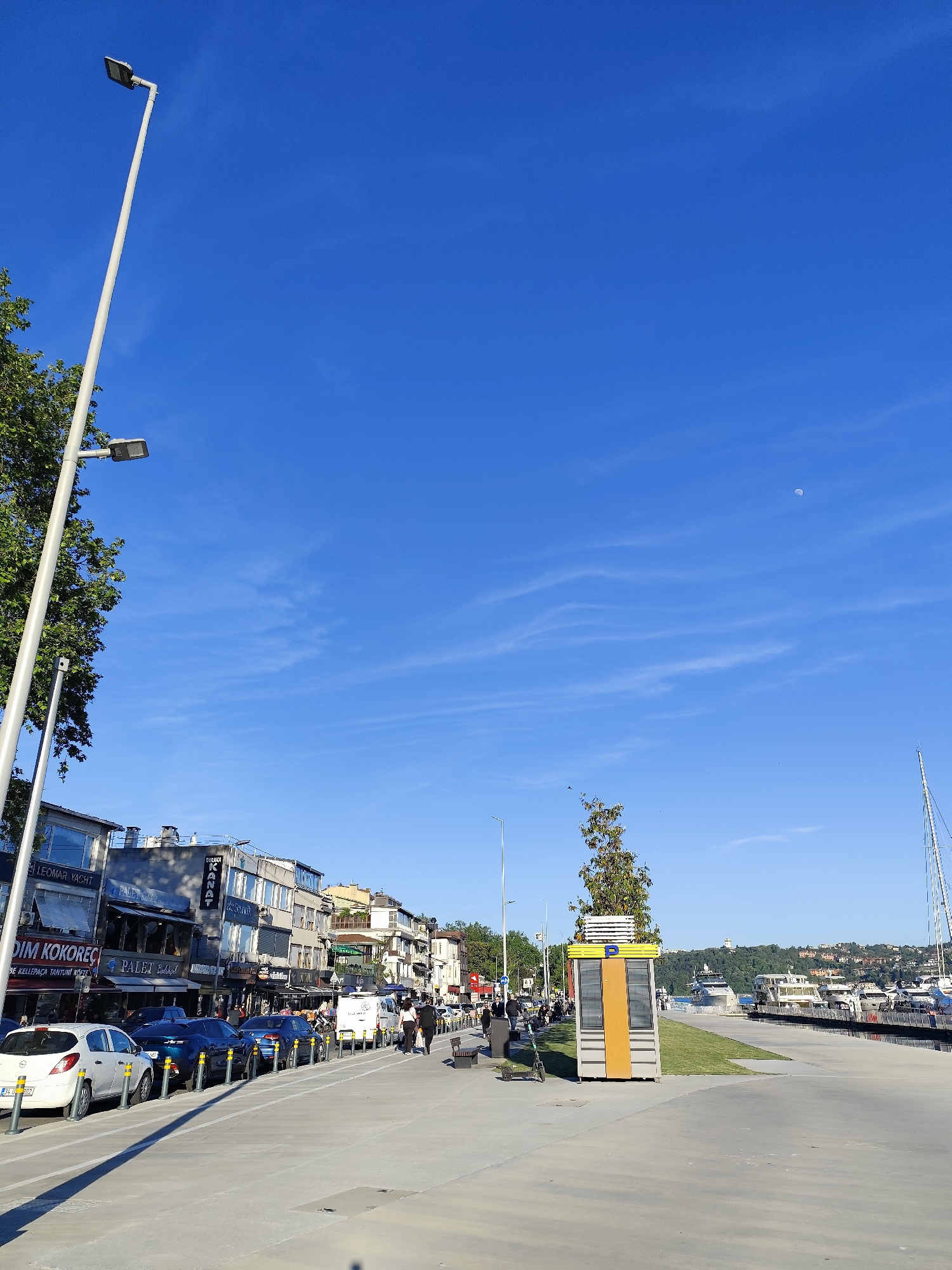 The image depicts a street scene in Sarıyer, Turkey, on a sunny day with a clear blue sky. The photograph is taken from a low angle, capturing the wide expanse of the sky, the street, and the buildings along it.
On the left side of the street, cars are parked. The buildings are mixed-use, featuring shops and restaurants on the ground floor and apartments or offices above. Signage indicates establishments like "LEOMAR YACHT," "PALET," and "DURAK KAFA." People are walking along the sidewalk, engaging in various activities.
On the right side of the street, there is a pedestrian walkway separated from the water's edge by a grassy area. A small yellow booth with a "P" symbol on top stands on the grassy area, possibly indicating a parking facility or tourist information. In the background, boats and yachts are docked, indicating a waterfront location. A hillside with houses and trees is visible across the water.
Additional elements in the scene include streetlights, trees, and a partial view of the sky where there is a crescent moon.