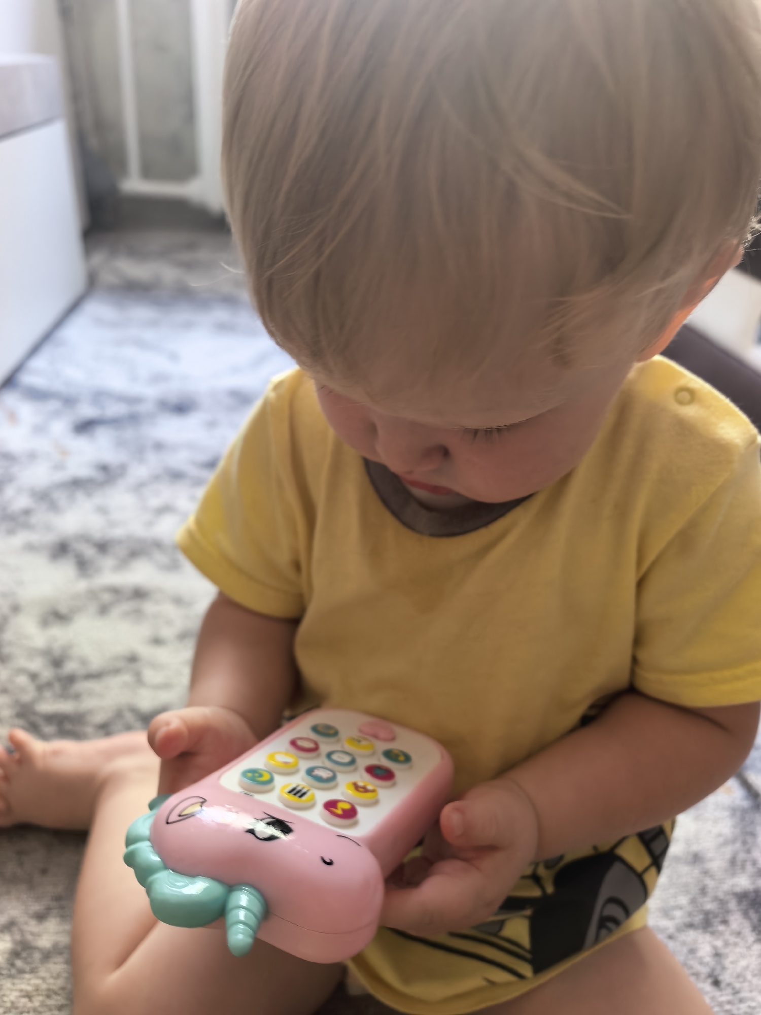 A blond-haired toddler is sitting on a patterned rug, holding a pink and turquoise toy phone. The toddler is wearing a yellow t-shirt. The background is blurred, but appears to be an indoor setting with furniture and part of a wall. The image does not contain enough information to determine the location within the city of Sterlitamak, Russia.