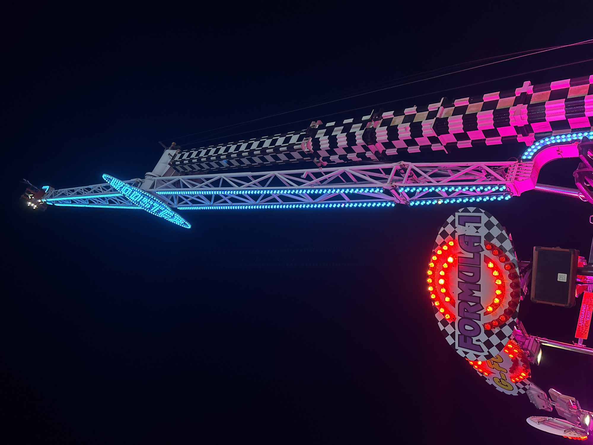 A low-angle shot of a carnival ride at night. The ride is decorated with lights. The ride's supports are illuminated with blue and pink lights. One arm of the ride has a checkered pattern, while another has a sign that reads "Formula 1". The background is a dark night sky. The location is potentially in Varna, Bulgaria, but this cannot be confirmed from the image.