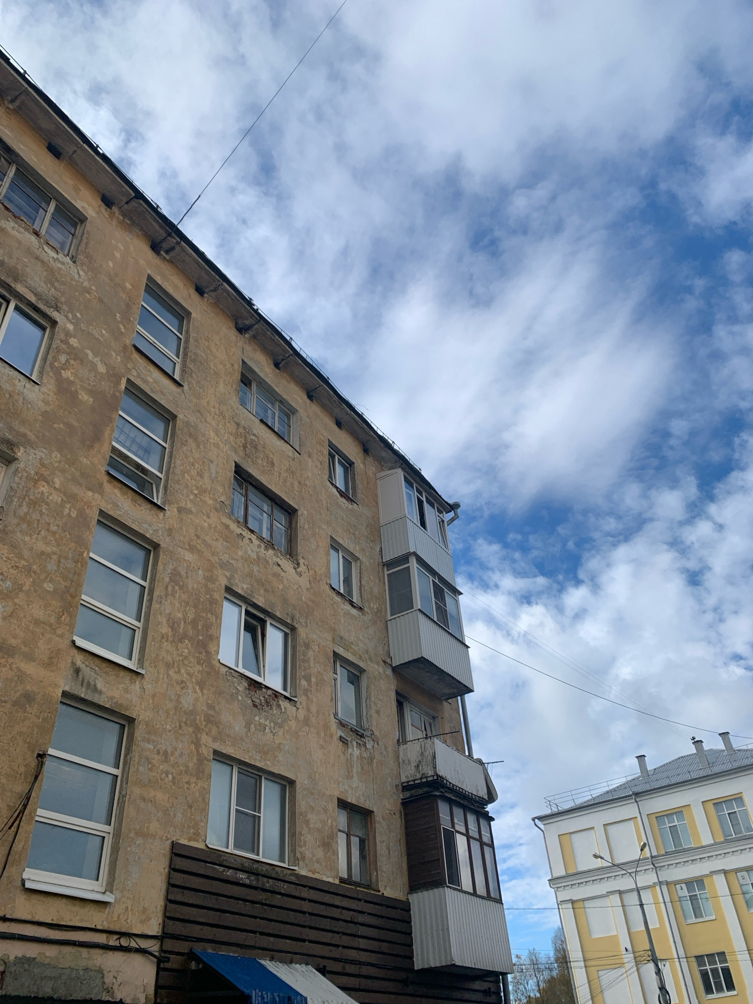 The image shows a view looking up at the side of a weathered, multi-story apartment building in Arkhangelsk, Russia. The building has a light brown, peeling facade and numerous windows, some with white frames and others with darker frames. There are balconies, one of which is enclosed with glass. A dark wooden fence-like structure is visible at the lower part of the building. In the background, to the right, is another building with a yellow and white facade and a grey roof. The sky is filled with white and grey clouds against a blue background. Utility wires are strung across the scene. There are no people visible in the image.