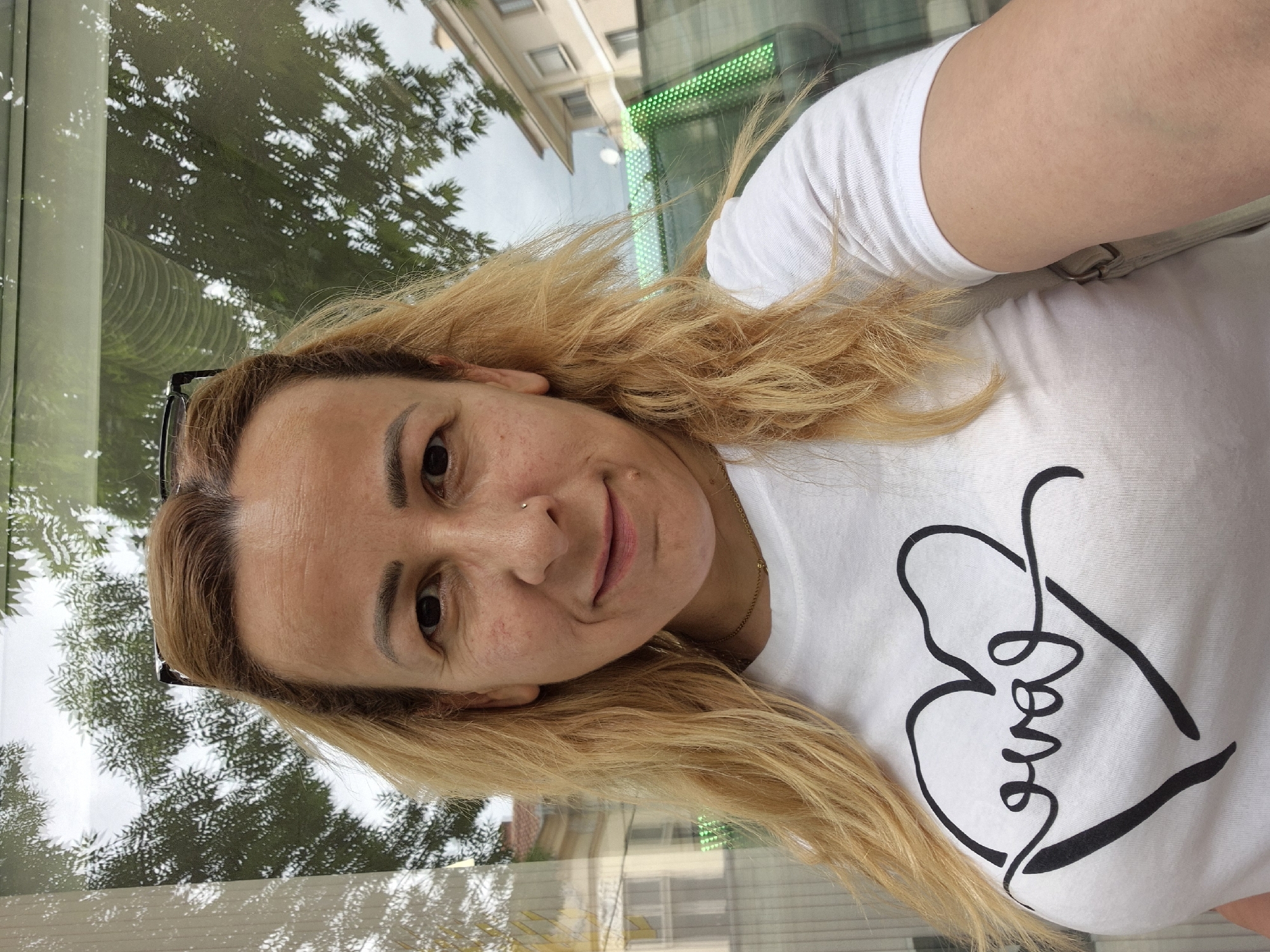 A blonde woman is posing for a selfie indoors, facing the camera at a slight angle. She has fair skin and is wearing a white t-shirt with a large black heart design incorporating the word "love." A small nose ring is visible. Her hair is blond and slightly wavy, falling over her shoulder. A pair of glasses are perched atop her head.
Behind her, a window reflects an exterior scene. Trees are visible, along with a building in the distance. The reflections suggest a daytime setting. Based on the green dot matrix on the building, the location might be in a city. The specific city and country are Unknown.