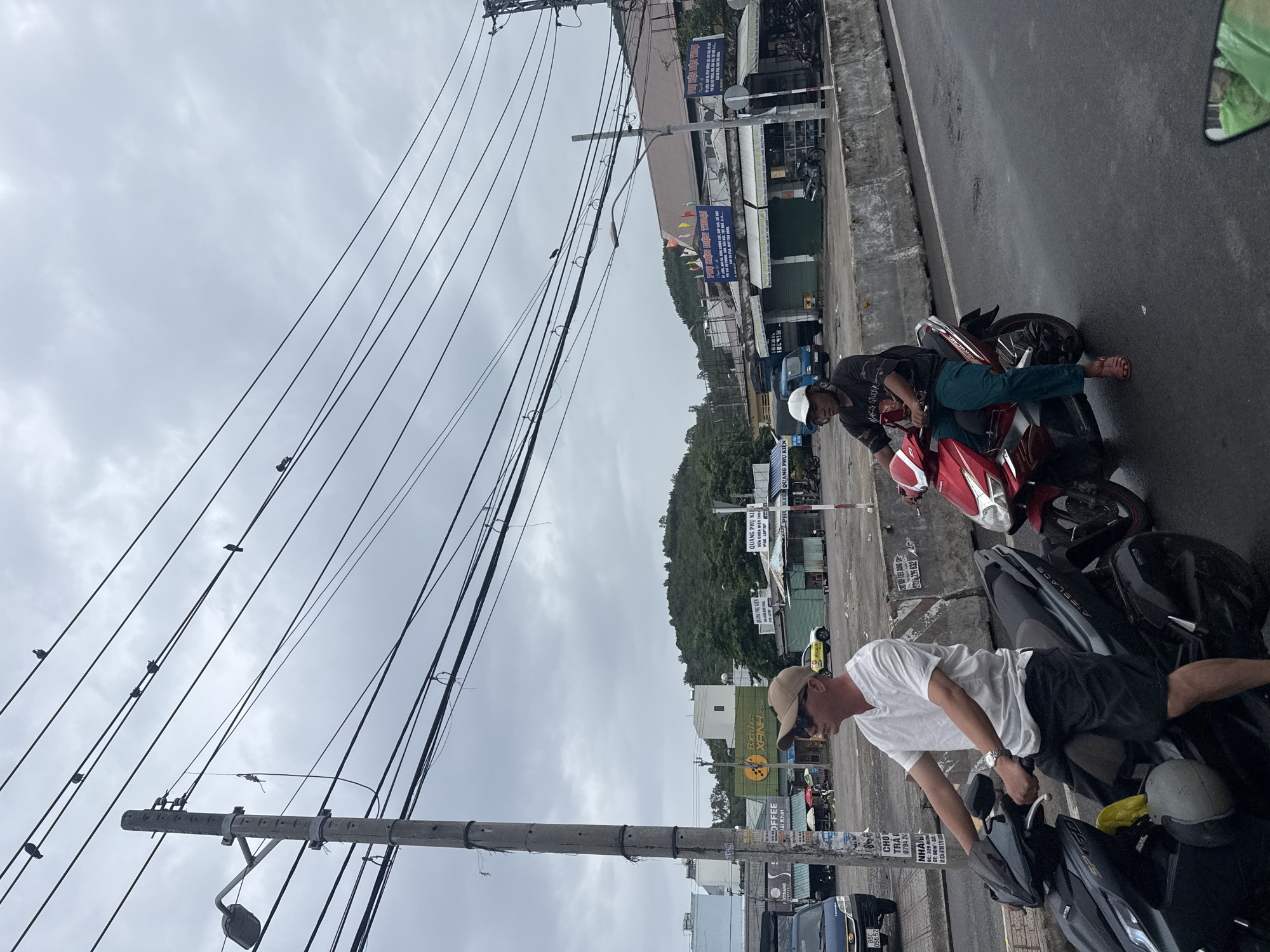 The image captures a street scene in Nha Trang, Vietnam, under a cloudy sky. The foreground features a man on a dark motorcycle, wearing a white t-shirt, dark shorts, sunglasses, and a tan baseball cap. He is looking to his right, away from the camera. To his right, a red motorcycle is ridden by another individual wearing a dark t-shirt, green pants, and a white cap, also looking forward. In the background, a street lined with shops and buildings is visible, with a prominent green building with the text "Bach Hoa Xanh" on it. Hills covered in greenery form the backdrop. Overhead, a dense network of electrical wires stretches across the frame, with several birds perched on them. The street appears wet, suggesting recent rain.
