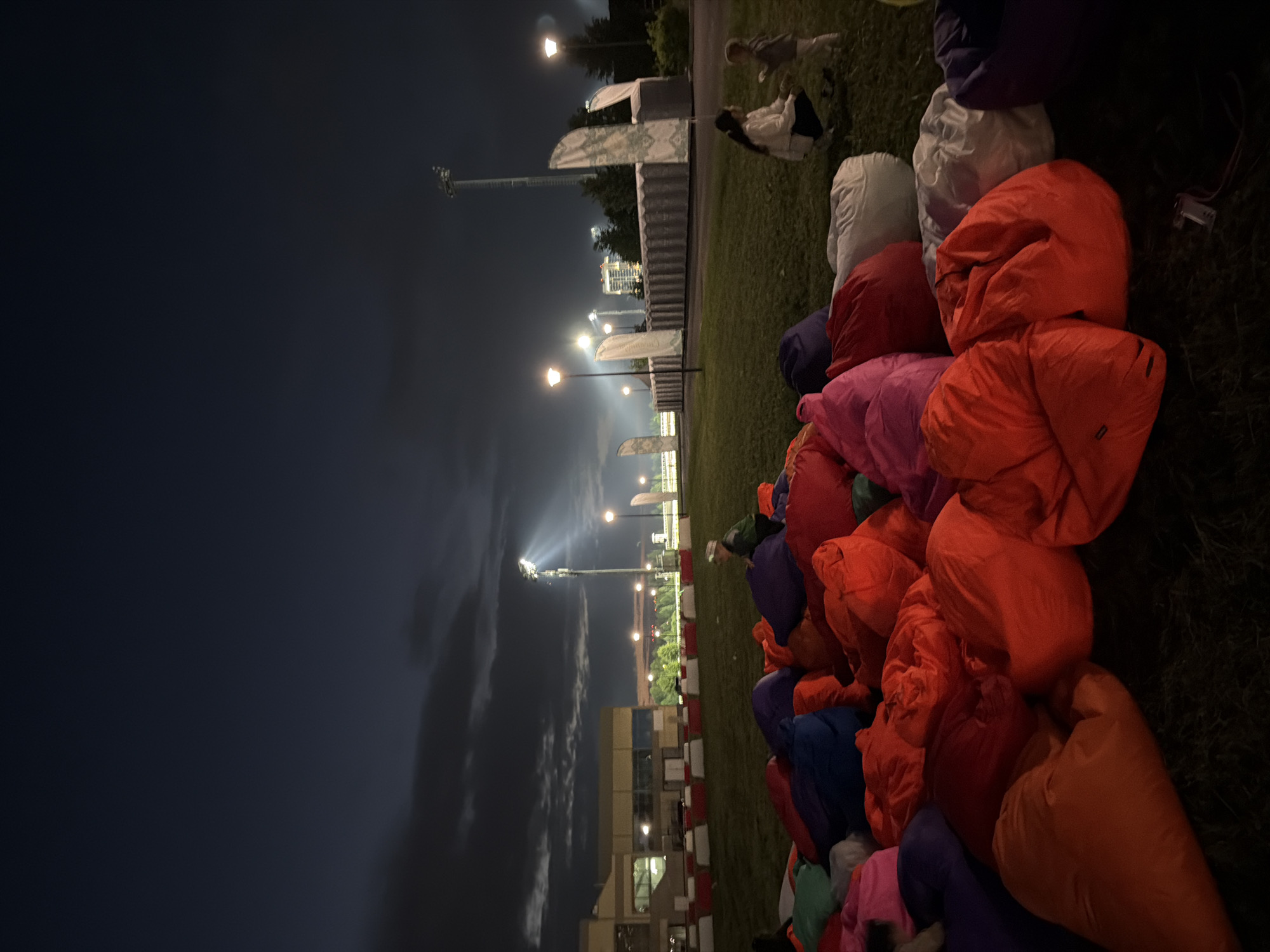 The image depicts an outdoor gathering at night, with a focus on a large collection of colorful beanbag chairs scattered across a grassy area. In the background, a modern building with large windows is visible, illuminated by artificial light. Several tall lampposts with spotlights are also present, casting beams of light into the dark sky, suggesting an event is or will be taking place. A few people are visible, including a woman and a young child interacting in the mid-ground, and another individual partially visible on a beanbag chair in the foreground. The overall scene suggests a relaxed, possibly social event like an outdoor movie screening or concert.