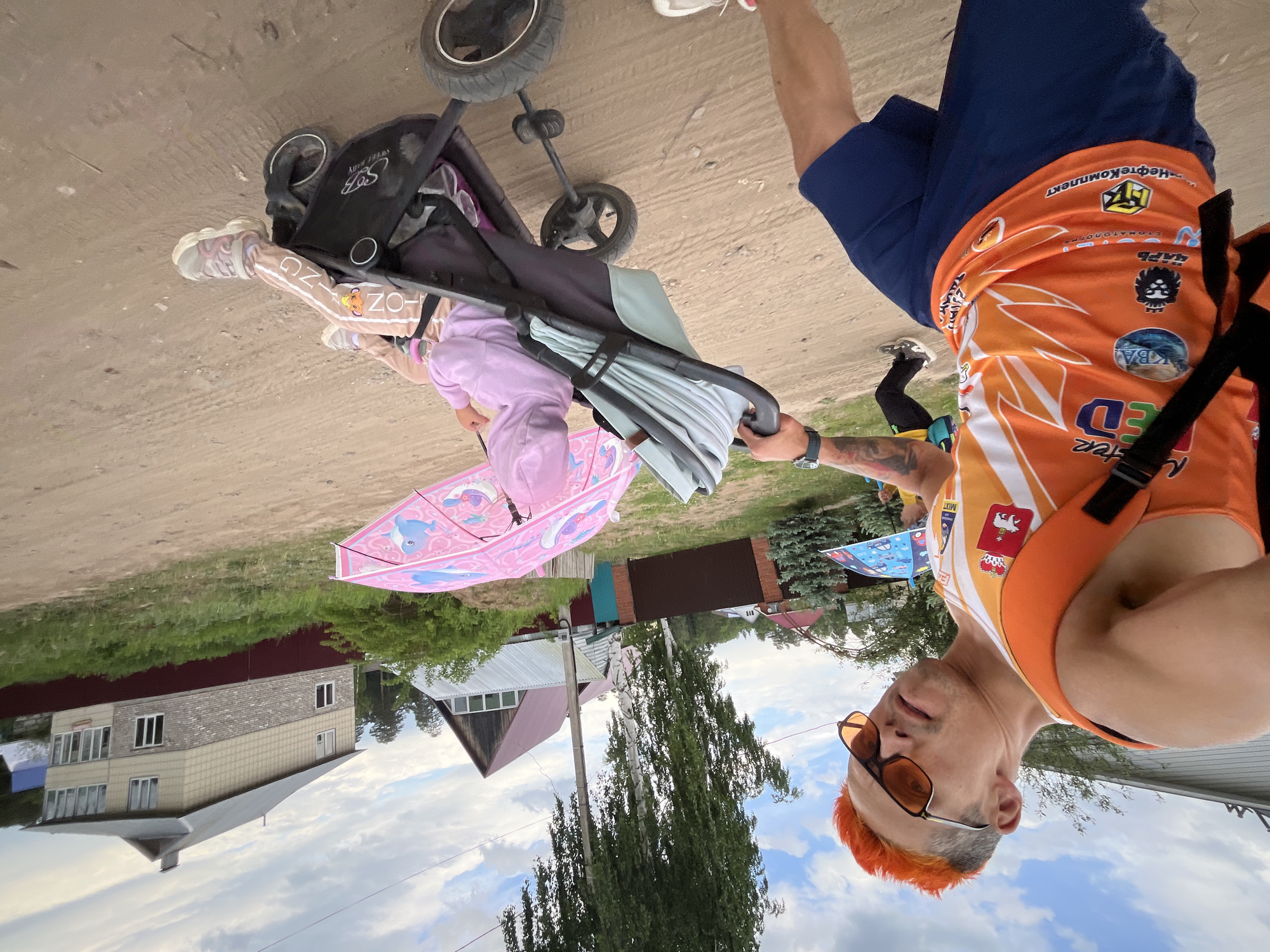 The image shows a man with orange hair and sunglasses wearing an orange shirt and blue shorts standing on a dirt path in what appears to be a suburban area. He is holding a baby stroller containing a young child under a pink umbrella. A house with a gray roof and cream-colored siding is visible in the background, along with trees and other greenery. Based on other clues, this photo was taken near Chaikovsky, Russia.