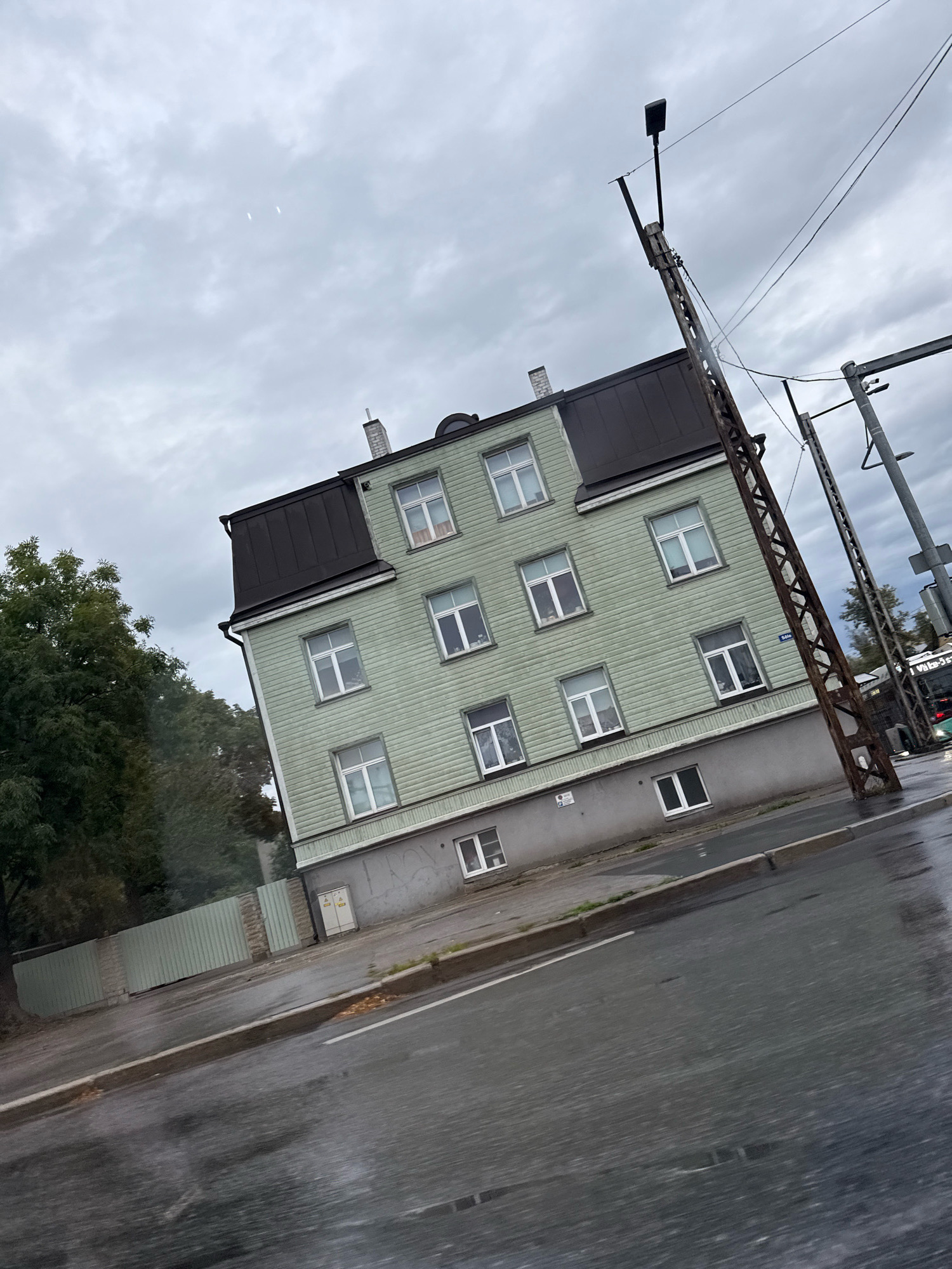 The image captures a street scene in Tallinn, Estonia, under a cloudy sky. A light green, multi-story building with a dark pitched roof dominates the left side of the frame. The building has numerous white-framed windows. A tall, rusty metal pole with electrical wires stretches upwards on the right side of the building. Adjacent to the building on the left is a dense green tree. The street in the foreground appears wet, indicating recent rain, with reflections visible on its surface. A light-colored fence runs along the side of the property. Further down the street, a bus with "Valga-Narva" written on its front is visible, along with other electrical poles and infrastructure. The overall impression is a somewhat overcast and damp urban environment.